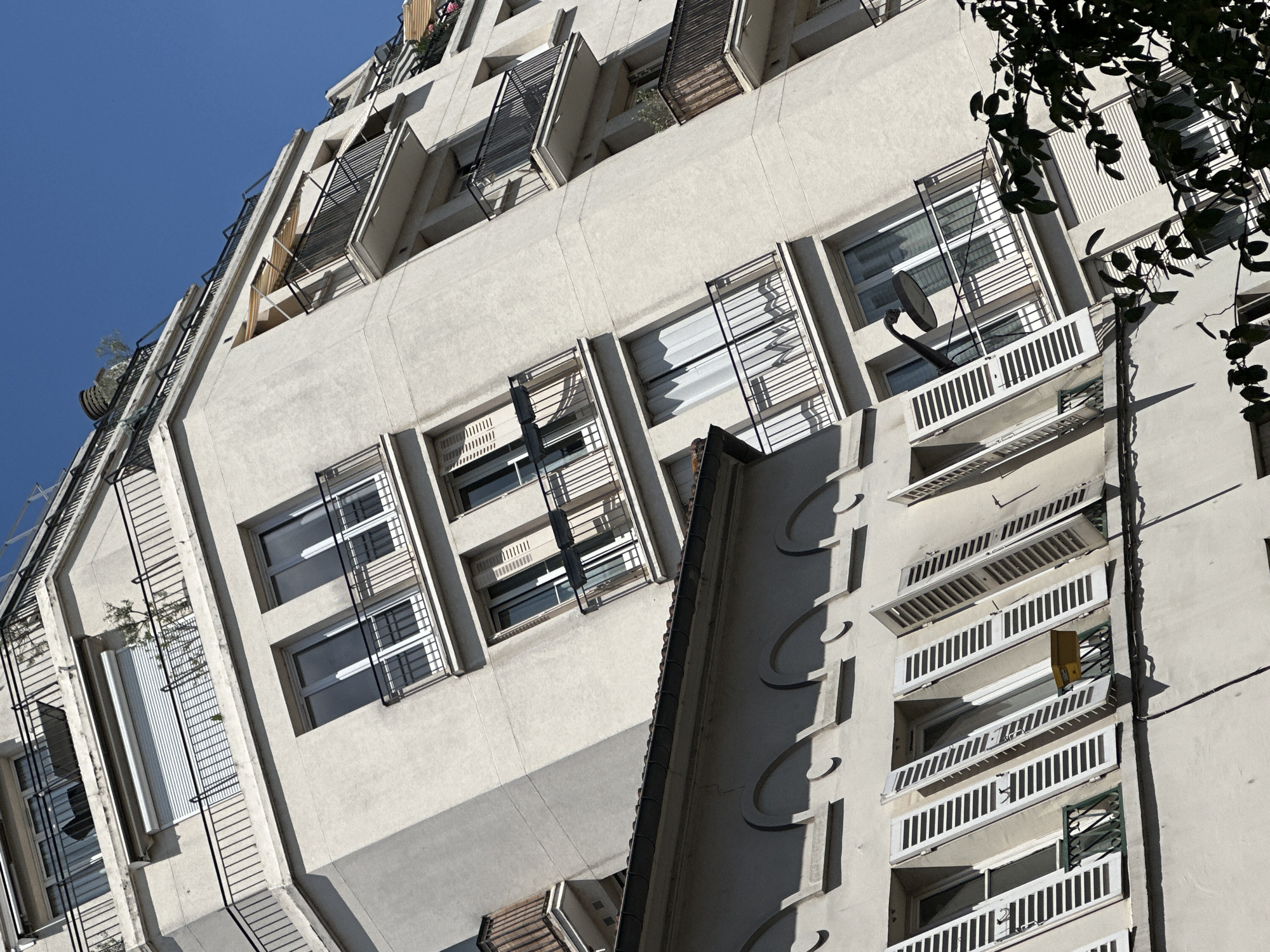 The image shows a close-up, low-angle view of the facade of a modern apartment building, likely in Paris, France. The building's exterior is a textured, light-colored concrete, with a series of recessed balconies and windows. Many of the balconies feature white slatted railings and some have louvred shutters, several of which are open. The windows are dark, reflecting the clear blue sky above.
There are no people visible in the image. The objects present include a satellite dish mounted on a balcony, some greenery on a balcony on the left side of the frame, and a small yellow object hanging from a railing on the right. The architecture suggests a mid-to-late 20th-century design, with a somewhat utilitarian but distinctive style, characterized by the repetitive geometric elements and the balconies.
The scene is set outdoors on a sunny day, indicated by the bright light and the clear blue sky. The strong shadows cast by the architectural elements and the tree branches in the upper right corner suggest it is midday. The overall impression is one of urban architecture bathed in sunlight. There is no visible text in the image.