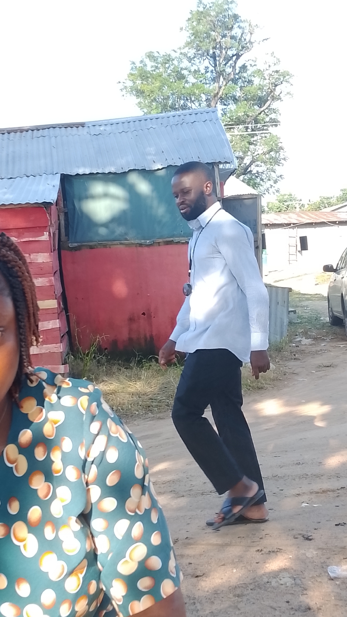 A man with dark skin and a beard is walking on a dirt road in Adankolo, Nigeria. He is wearing a light blue striped long-sleeved shirt and black pants, with black sandals. He has a black lanyard with a circular object hanging from it around his neck. To the left of the frame, the shoulder and upper chest of a woman wearing a teal top with an abstract brown and white pattern are visible. Behind the man, there is a small red building with a corrugated metal roof and a green tarpaulin covering an opening. Further in the background, there are more buildings with corrugated metal roofs, a tree with lush green foliage, and a silver car parked on the right side of the road. The ground is covered in dirt and sparse dry grass. The lighting suggests it is daytime, with bright sunlight casting shadows.