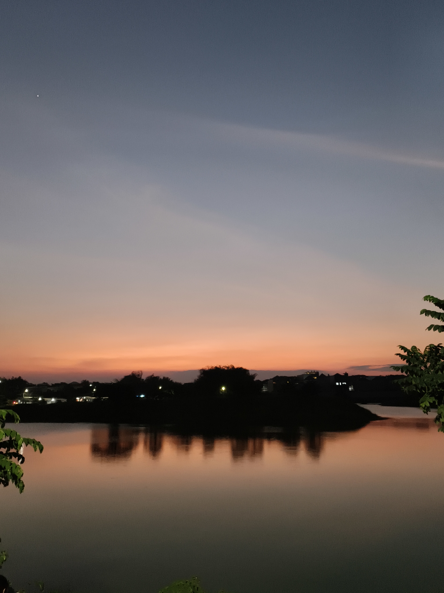A serene sunset scene unfolds over the city of Surabaya, Indonesia, as viewed from a vantage point near the Kenjeran Lama Beach area. The sky is a gradient of soft blues and oranges, with a single bright star visible in the upper left quadrant. A silhouetted landmass, dotted with the faint lights of buildings, stretches across the middle ground, reflected in the calm waters of a lake or bay in the foreground. The water mirrors the colors of the sky and the dark shapes of the trees and structures on the shore, creating a peaceful, almost ethereal atmosphere. Lush green foliage frames the right side of the image, adding a touch of nature to the urban landscape. The overall impression is one of tranquility and natural beauty, despite the presence of the city.