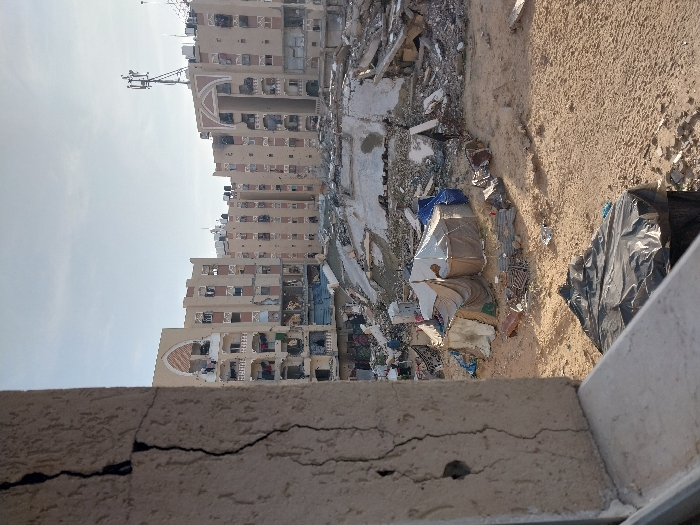 The image captures a scene of urban destruction in Khan Yunis, Palestinian Territory. In the immediate left foreground, a light-colored concrete or stucco pillar, likely part of a building's structure, displays a prominent, dark vertical crack running its length and a small circular indentation. The midground consists of a large, flat expanse of light brown sand and dirt, interspersed with scattered debris including concrete rubble and fragmented materials. Clustered towards the left-center midground are several makeshift shelters constructed from various materials, including beige canvas, blue tarpaulins, and possibly blankets or corrugated sheets. A lone individual, clad in dark clothing, stands in the sandy area to the right of these shelters, near a mound of rubble. The background features a complex of multi-story residential buildings, predominantly light brown, exhibiting extensive damage; many windows are shattered or missing, and sections of the structures appear partially collapsed or heavily impacted. Architectural elements like rounded arches are visible on the facades. A cellular communication tower with multiple antennas is mounted on the roof of one of the background buildings. The sky above is clear and light blue, indicating daylight.