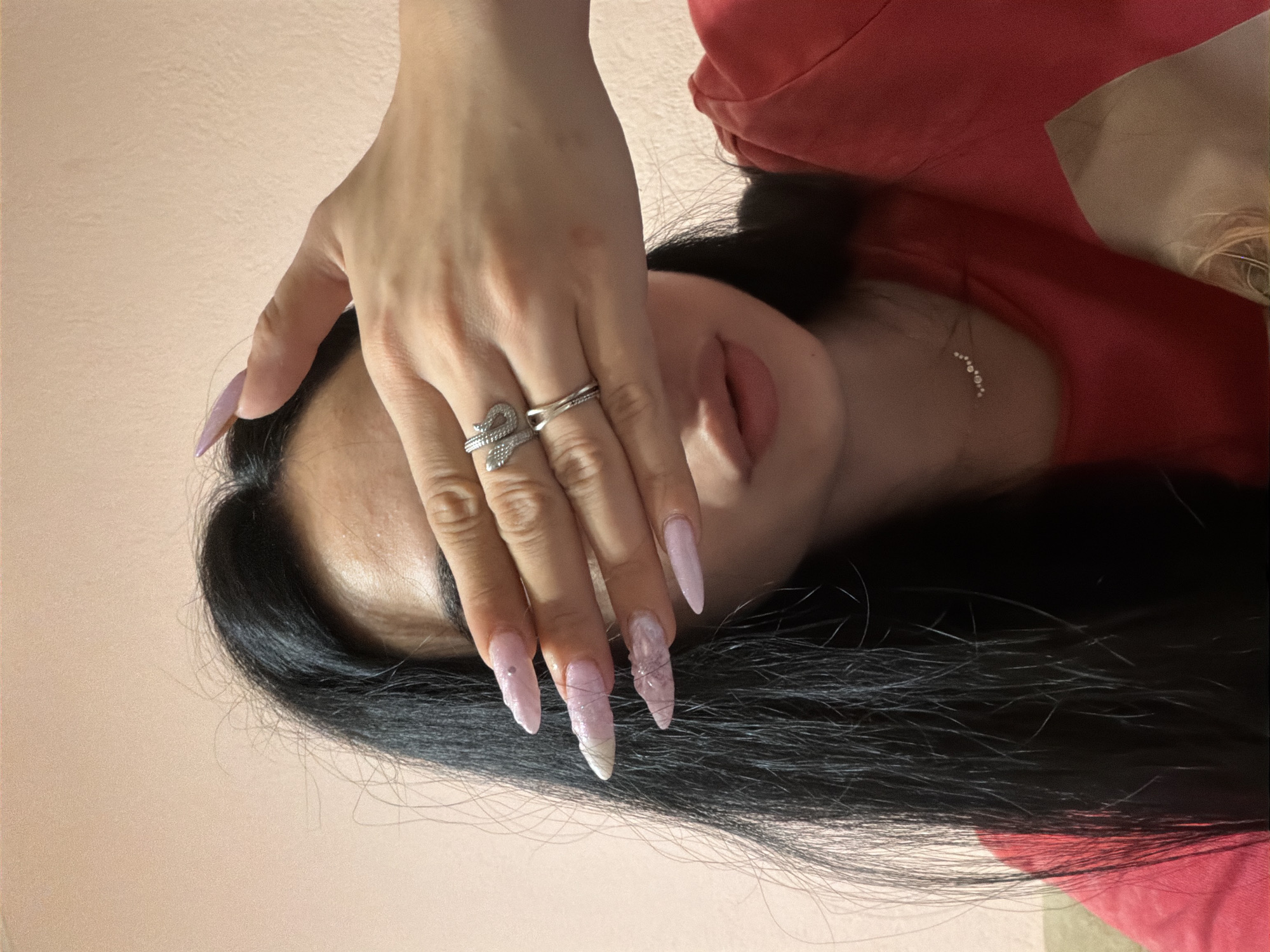 The image features a close-up view of a person's hand, adorned with elaborate light pink, stiletto-shaped acrylic nails, some embellished with glitter and what appear to be tiny embedded threads or fibers. Two silver rings are visible on the fingers, one a simple band and the other a decorative piece resembling a coiled snake. The hand is partially obscuring the person's face, revealing their lips and a hint of their chin. They have long, dark, straight hair that cascades around their head and shoulders. The person is wearing a red garment, possibly a shirt or dress. A subtle necklace with small, circular charms is visible on their neck. The background is a soft, neutral pink, suggesting an indoor setting. The lighting is soft, and the overall mood conveyed is one of personal adornment and style. There is no visible text in the image. The provided location context is Stara Zagora, Bulgaria.