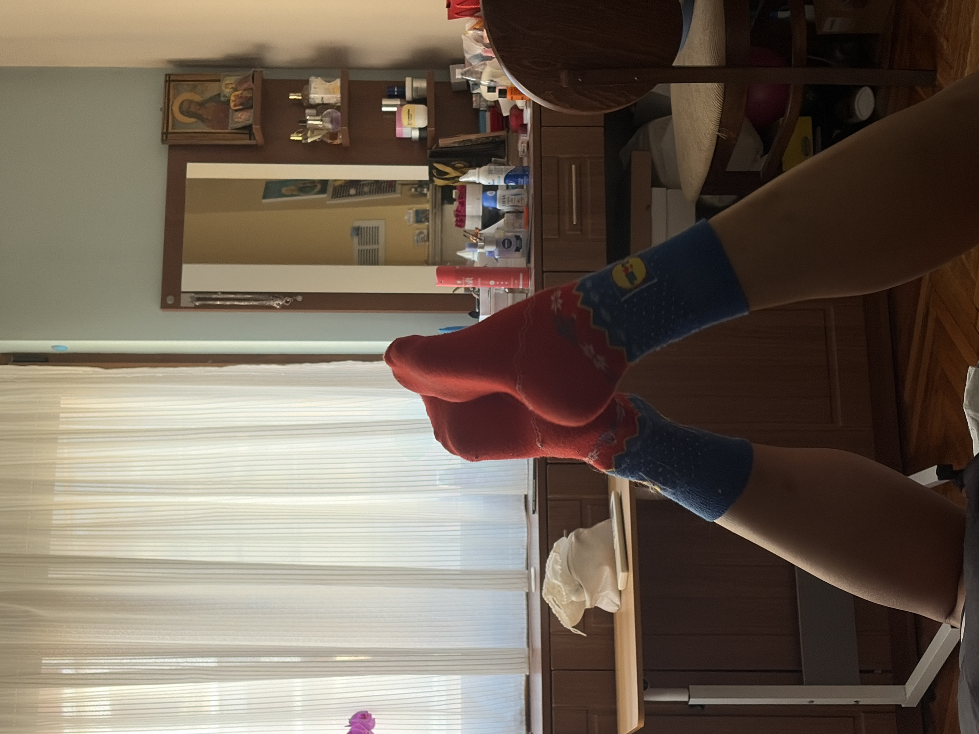 The image depicts a person's legs, clad in red and blue socks with a patterned design, extended upwards. The legs are positioned in front of a wooden dresser and a white curtain. On the dresser, various items are visible, including bottles of toiletries, a mirror with religious icons and a necklace on a shelf, and a remote control on a small table. A wooden chair with a woven seat is situated to the right of the dresser. The scene appears to be an indoor setting, likely a bedroom, and there is no discernible connection to the city of Vratsa, Bulgaria.