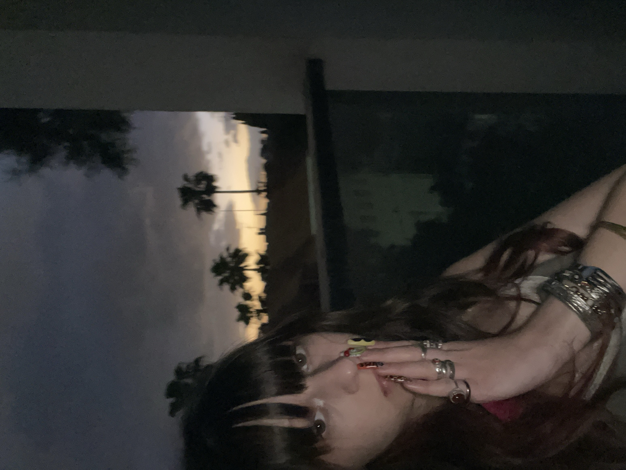 A person with dark hair and bangs, adorned with rings and a bracelet, is posing in front of a sunset scene with palm trees. It is dusk. Behind the person are exterior features of a structure, possibly a building. Given the landscape, this may potentially be somewhere in Raanana, Israel, a city characterized by palm tree lined streets. Further location detail is indeterminate from the current frame.