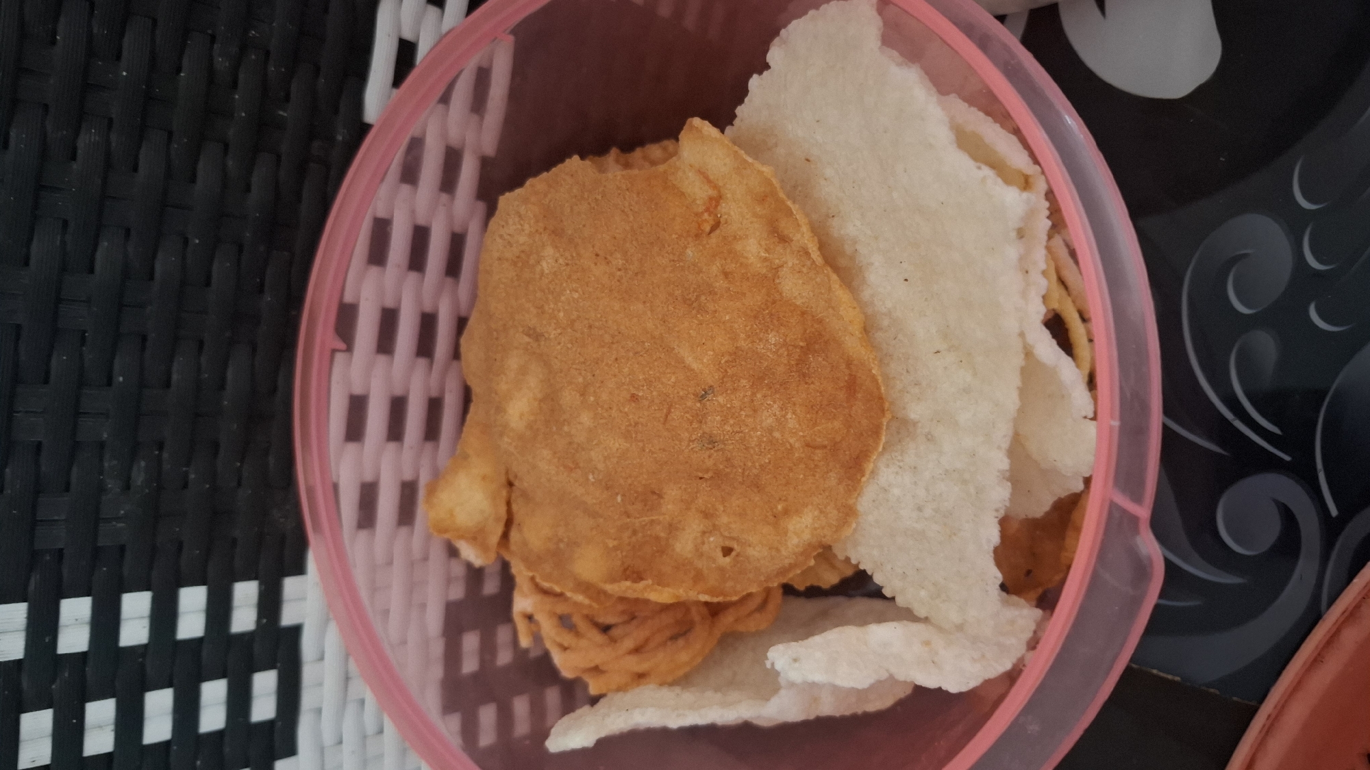 A close-up, high-angle shot shows a pink plastic bowl filled with various crackers. The crackers are layered and include a large, round, orange cracker with a slightly uneven surface in the center. Beneath it, several white, translucent crackers are visible. To the left and behind the orange cracker, a pile of thin, orange, string-like crackers is present. The background features a black and white woven surface, suggesting a textured mat or upholstery. The bowl rests on a black surface with a white swirling pattern. The image is likely taken in Ciharashas, Indonesia, as indicated by the context provided.