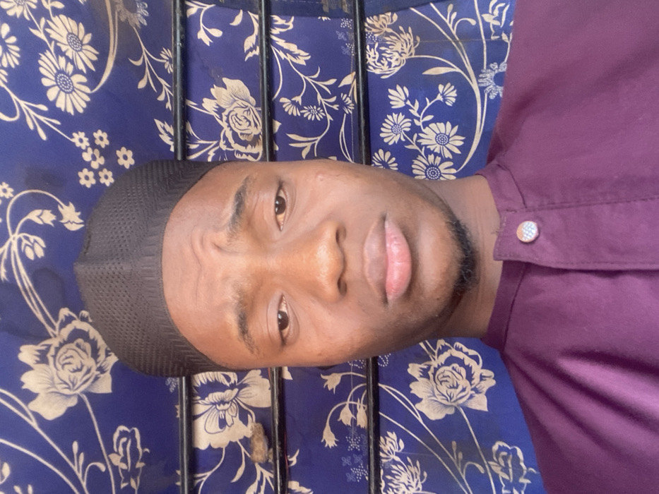 A young adult male is depicted in a close-up, head-and-shoulders portrait shot within an indoor setting in Katcha, Nigeria. He wears a black, mesh-textured kufi cap and a deep purple collared garment with a single circular, silver-toned decorative button visible at the neck closure. His skin tone is dark. His eyebrows are slightly furrowed, his eyes are open and directed forward, and his mouth is closed in a neutral expression. A faint beard and mustache are present. The background consists of a blue fabric featuring a repetitive pattern of light-colored floral and leaf motifs. Two horizontal dark metal bars are visible across the background, and a small, irregular brown object rests on the left side, positioned between the bars.