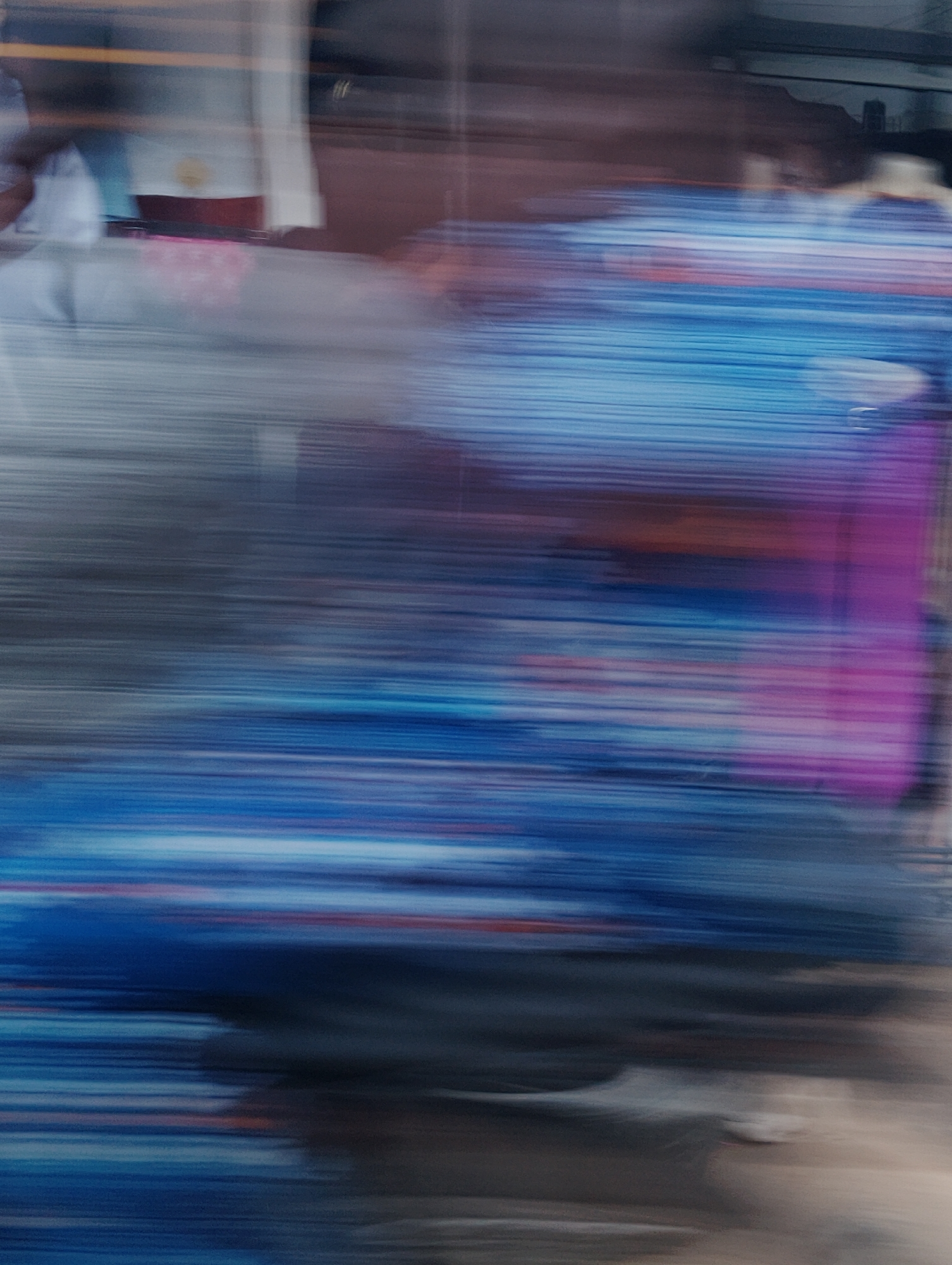 The image captures a blurred, streaky view of what appears to be a market scene in Isheri, Nigeria. The predominant colors are various shades of blue, with streaks of pink, red, and grey intermingling. The blur suggests motion, possibly due to the photographer or subjects moving, or a slow shutter speed. In the background, there are hints of structures and possibly people, but they are indistinct. The overall impression is one of movement and the vibrant, fluid nature of a busy urban environment.