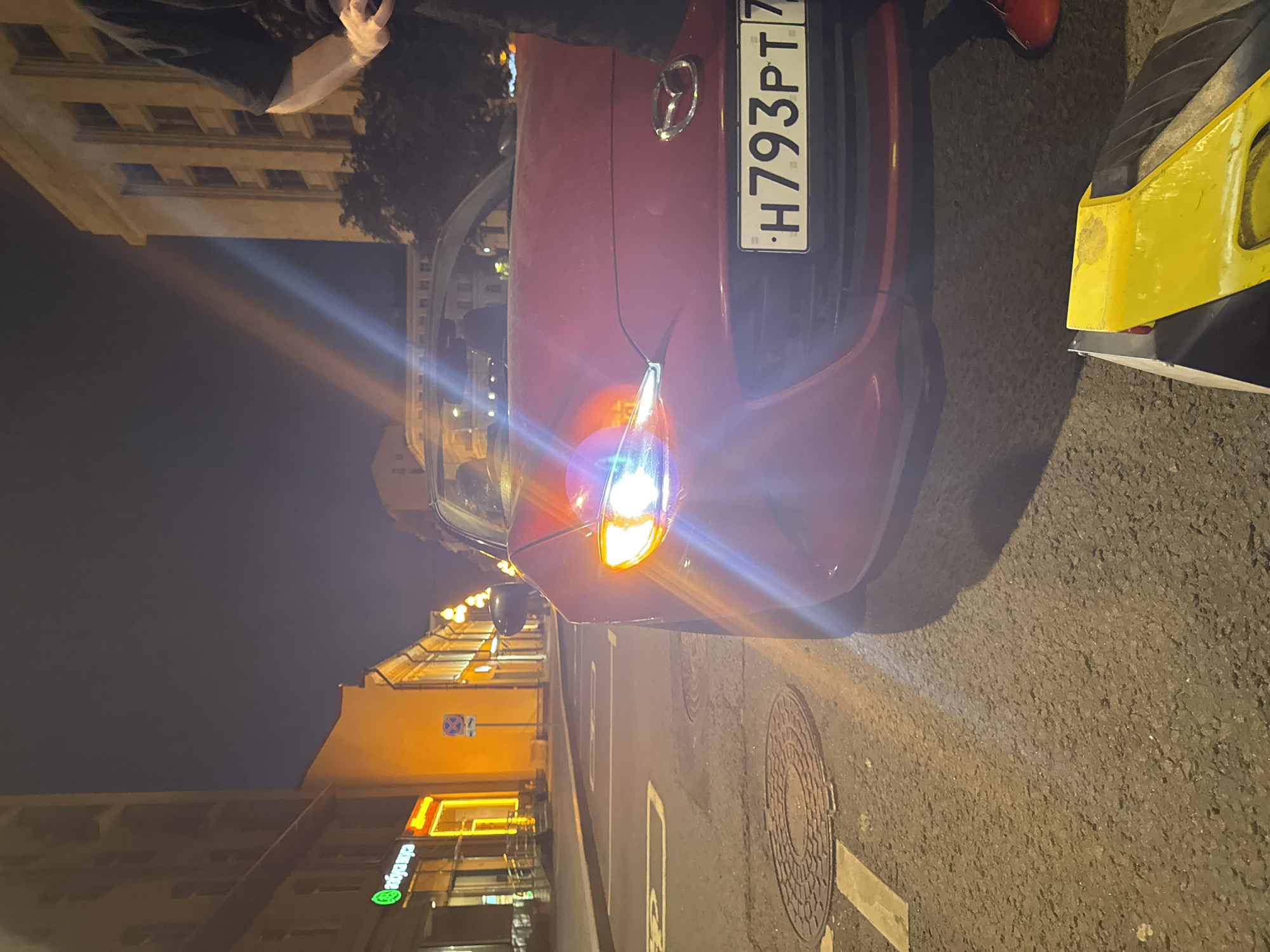 A red Mazda convertible is parked at night on a city street. The car's headlights are on. A person's legs, wearing red shoes, are visible on the right side of the frame. In the foreground, a yellow object, possibly a piece of equipment or a bag, is on the ground. The street is paved and has a manhole cover. Buildings line the street on both sides, and streetlights are visible in the background, illuminating the scene. The license plate on the car reads "H793PT77". The building on the left has a sign with green and white lettering, possibly in Russian. The overall impression is a nighttime street scene in an urban environment.