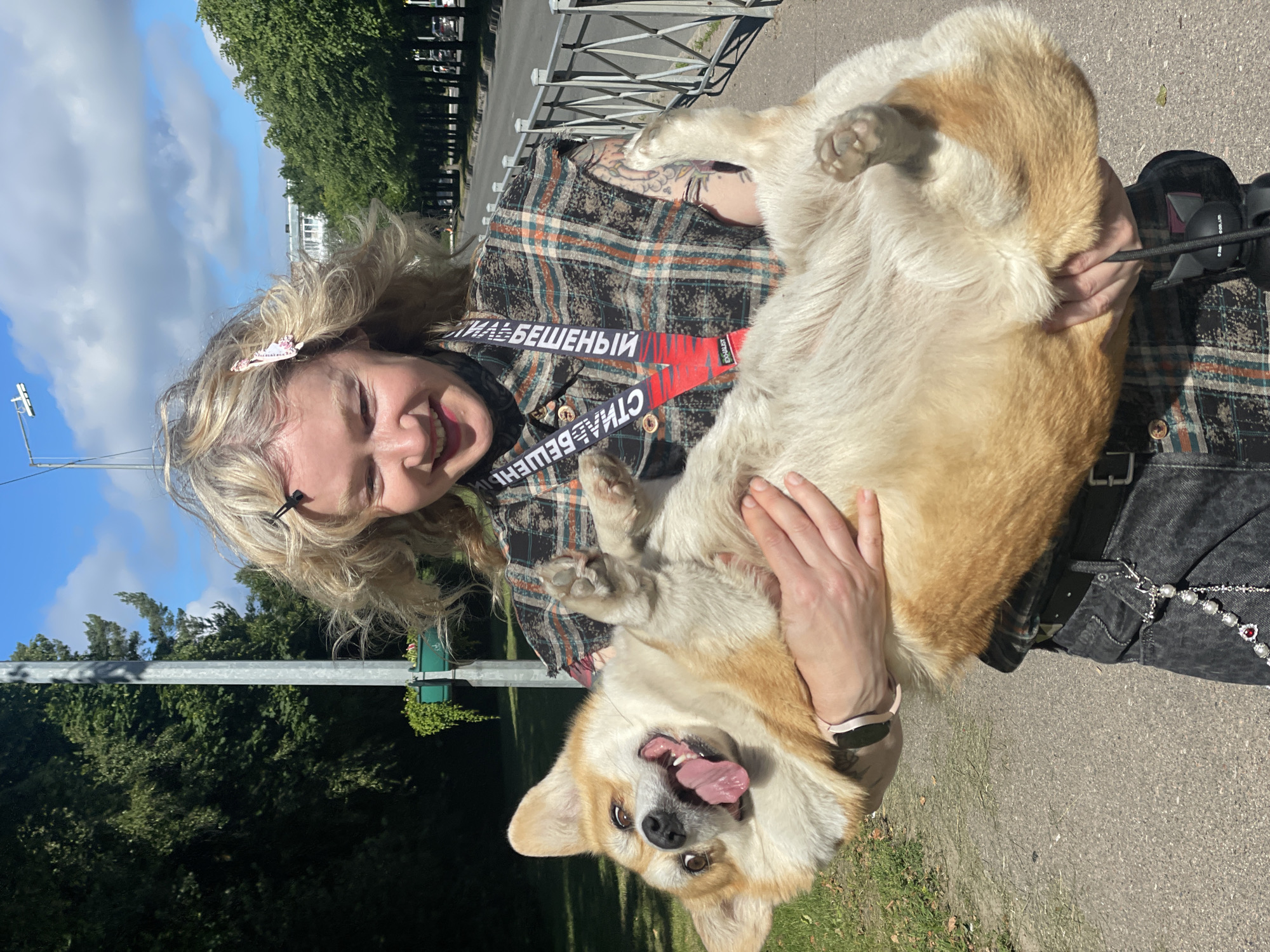 The media file shows a woman holding a corgi dog outdoors. The woman has blonde, wavy hair and is smiling widely, with her eyes closed. She is wearing a plaid shirt and has tattoos on her arm. A red strap with text is visible across her chest. The corgi dog is light brown and white, with its tongue hanging out and its mouth open in a happy expression. It is being held upside down by the woman, with its paws in the air.
The scene appears to be a park or outdoor recreational area, with trees and a paved path visible in the background. The sky is blue with some clouds, suggesting it is daytime and likely pleasant weather. A metal fence and a lamppost are also present. A small green planter with ivy is attached to the lamppost.
The visible text on the red strap appears to be in Cyrillic and says "СТИЛЬ БЕШЕНЫЙ", which translates to "MAD STYLE". Another word, "EXALIST", is visible in smaller font.
The overall mood of the image is joyful and playful, captured in the smiling faces of both the woman and the dog, and the dog's energetic pose. The setting suggests a casual outing on a sunny day.