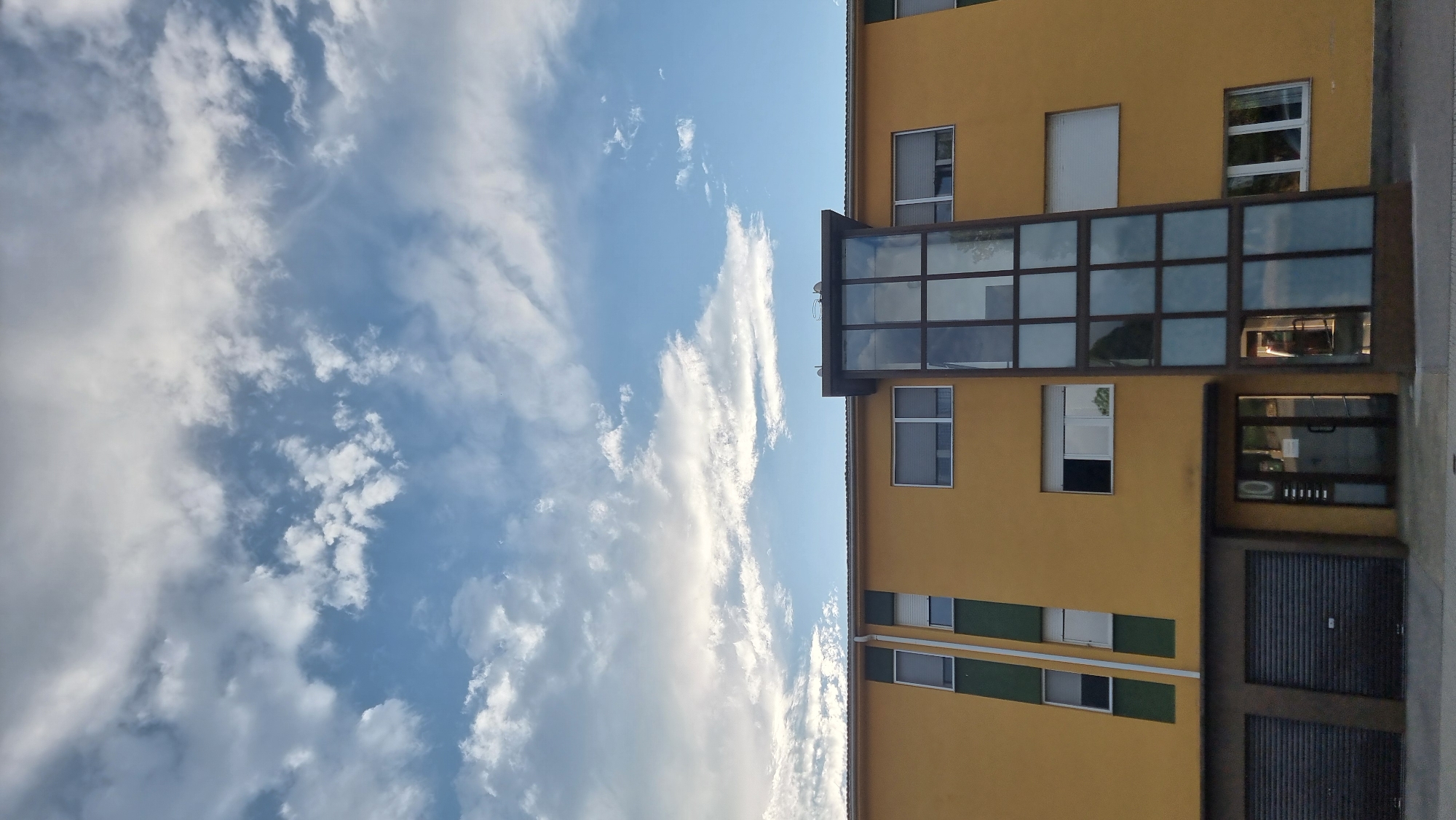 The image shows the exterior of a yellow, multi-story building in the city of Sant Joan Les Fonts, Spain. The building features several windows with blinds and a modern glass elevator shaft. On the ground floor, there are two dark roller doors, a glass entrance door with the number "10" above it, and another glass-fronted area to the right of the entrance. The sky above is filled with a mix of white and gray clouds against a blue backdrop.