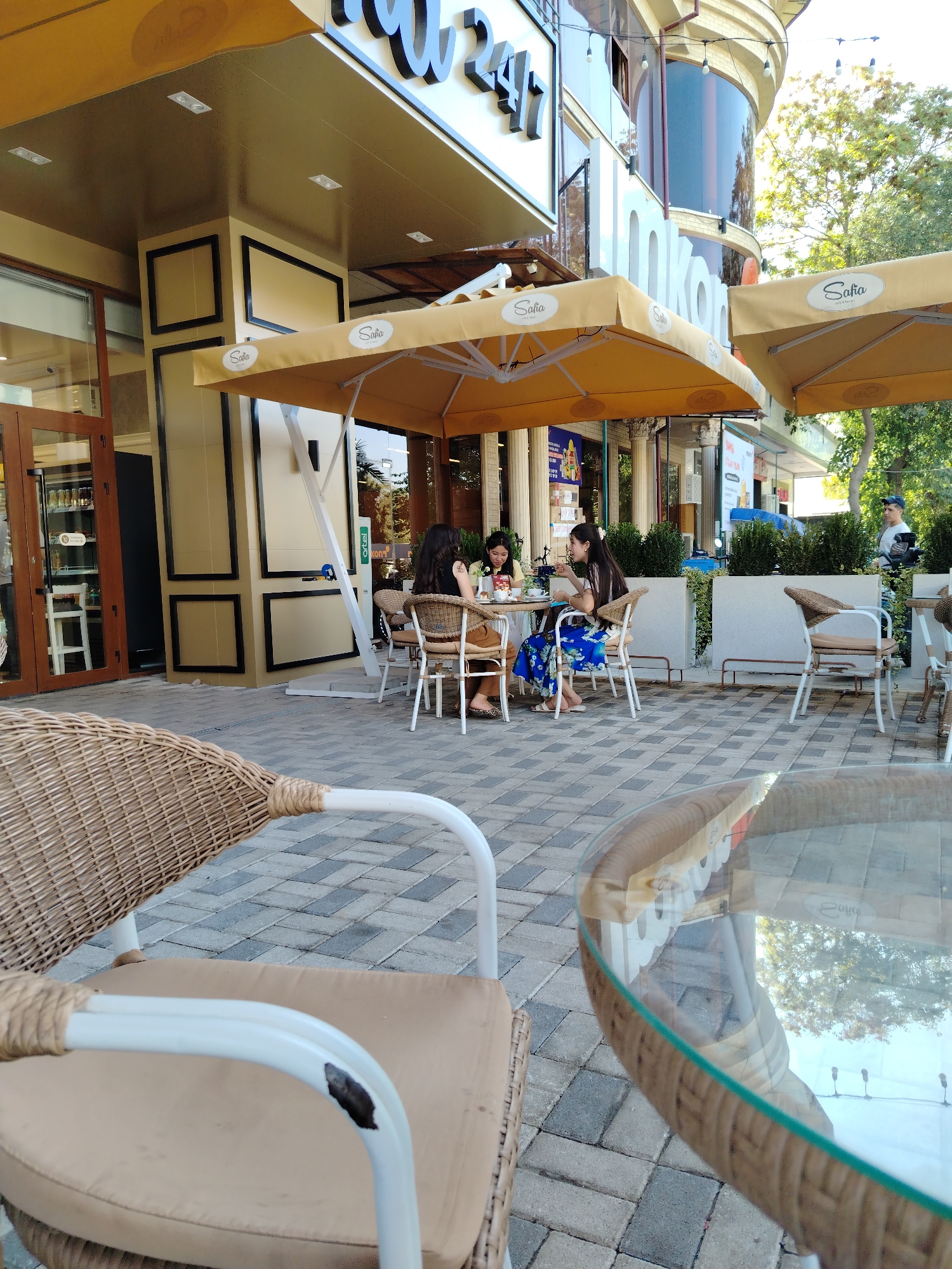 The image depicts the outdoor seating area of a cafe or restaurant located on the street in the city of Almalyk, Uzbekistan. In the foreground, a wicker chair with a tan cushion is partially visible. Further back, a round glass-top table with a wicker base is present, reflecting the surroundings. At a table occupied by three individuals, two women are seated, and one is standing. They are engaged in conversation and appear to be dining. Another table with chairs is set up nearby. The cafe is shaded by yellow awnings. The ground is paved with interlocking gray and brown bricks. The facade of the establishment features a beige exterior with dark framed rectangular elements. Trees and the sky are visible in the background.