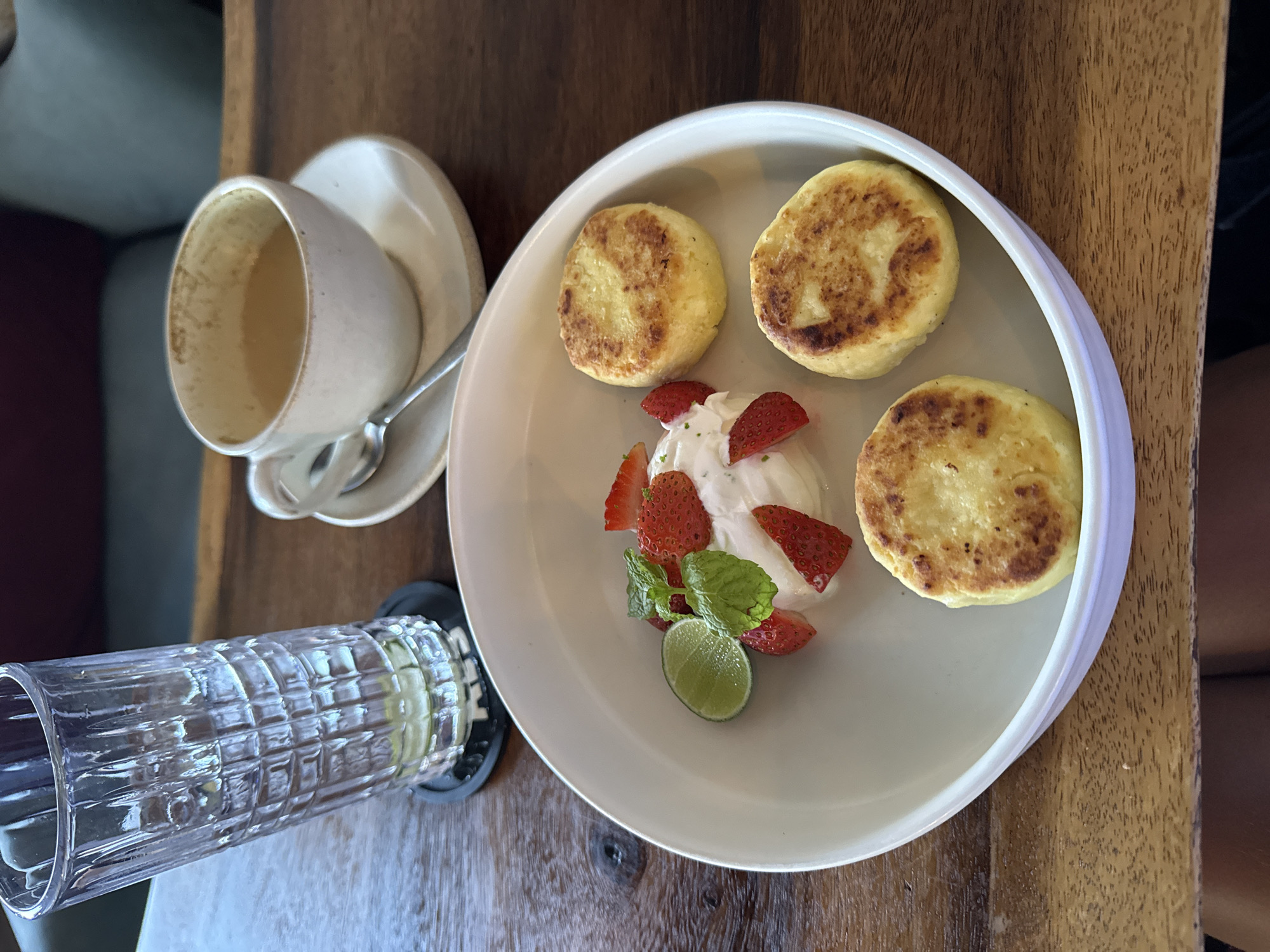 On a wooden table, a white plate holds three golden-brown syrniki (small cheese pancakes) arranged around a dollop of white cream topped with sliced strawberries, mint sprigs, and a lime wedge. Next to the plate is a white cup and saucer with a spoon, containing a light-colored beverage. Adjacent to the plate is a textured glass filled with a yellow liquid. The scene is likely set in a cafe in Canggu, Indonesia.
