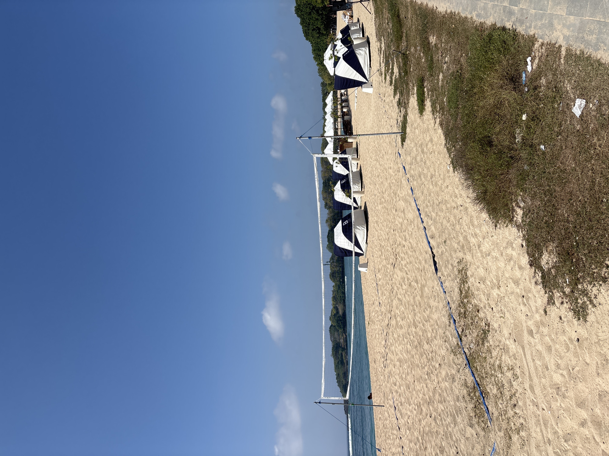 The image depicts a sandy beach in Kuta Selatan, Indonesia, under a clear blue sky with scattered white clouds. A volleyball net is set up on the beach, with the ocean and a tree-covered island visible in the background. Several white and navy blue striped tents or cabanas are lined up on the sand, some with white awnings extended. A paved walkway runs along the edge of the beach, bordered by overgrown grass with scattered litter. A single person, partially visible, is sitting on the sand near the volleyball net.