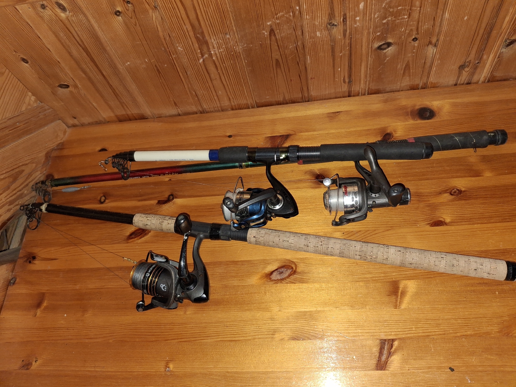This image depicts a collection of three fishing rods with reels, vertically suspended against a wooden wall, likely in a storage area. The rods are arranged in a slight diagonal line. The leftmost rod is predominantly cork-handled with a black and silver reel. The middle rod appears to be a darker color with a blue and silver reel. The rightmost rod is black with a white section near the top and a black reel. The wooden wall exhibits a visible grain pattern and natural knots. The lighting suggests an indoor environment. The scene is static, with no people or action present. The location is identified as Åsgårdstrand, Norway.