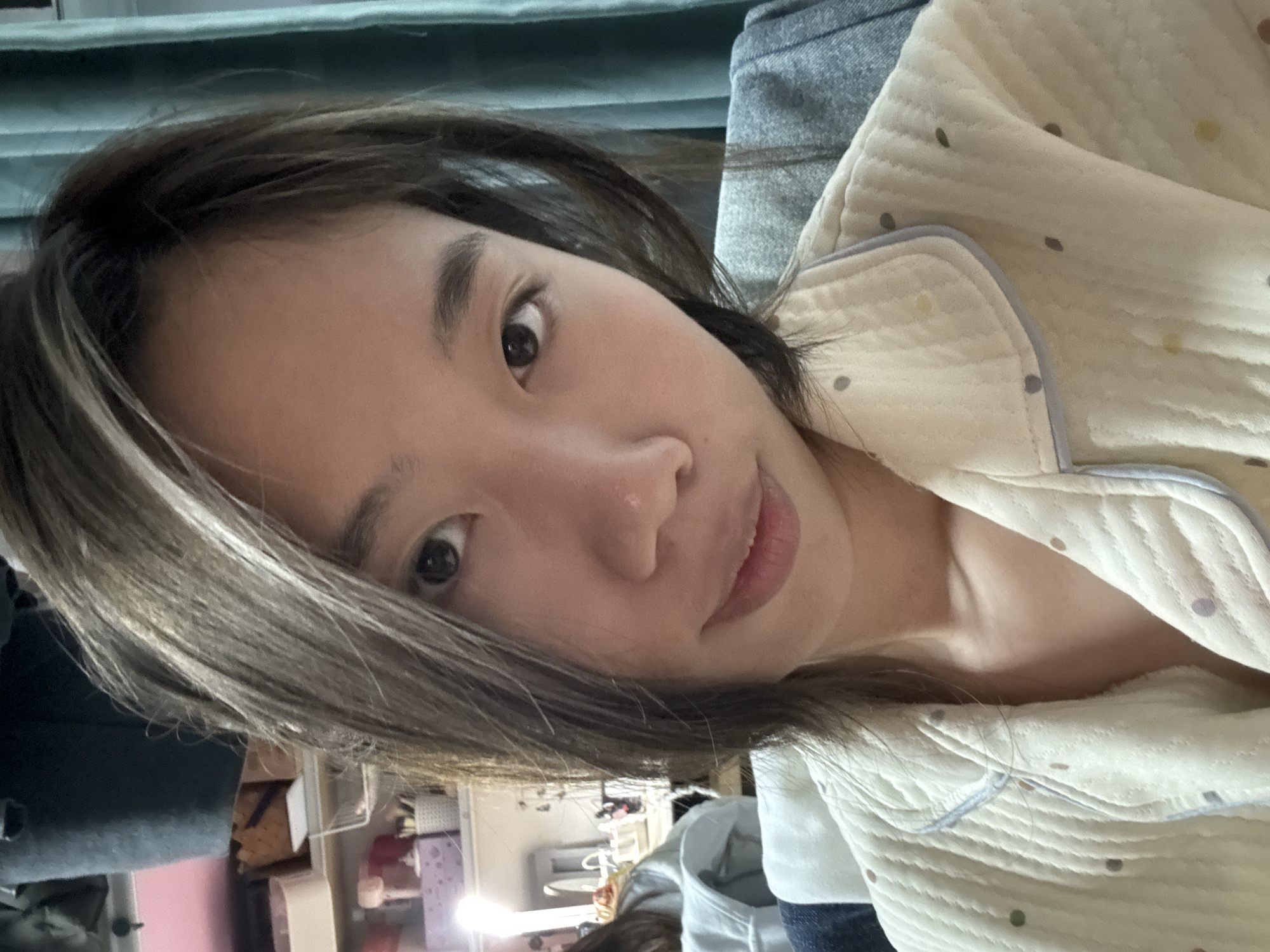 A close-up portrait shows a young Asian woman with dark hair and light skin. She is wearing a light-colored, quilted pajama top with colored polka dots and light blue trim. The background is partially visible, showing a blue curtain and a messy room with a white desk and various items on shelves. There is no indication of the location being in the city of Nan'an District, China.