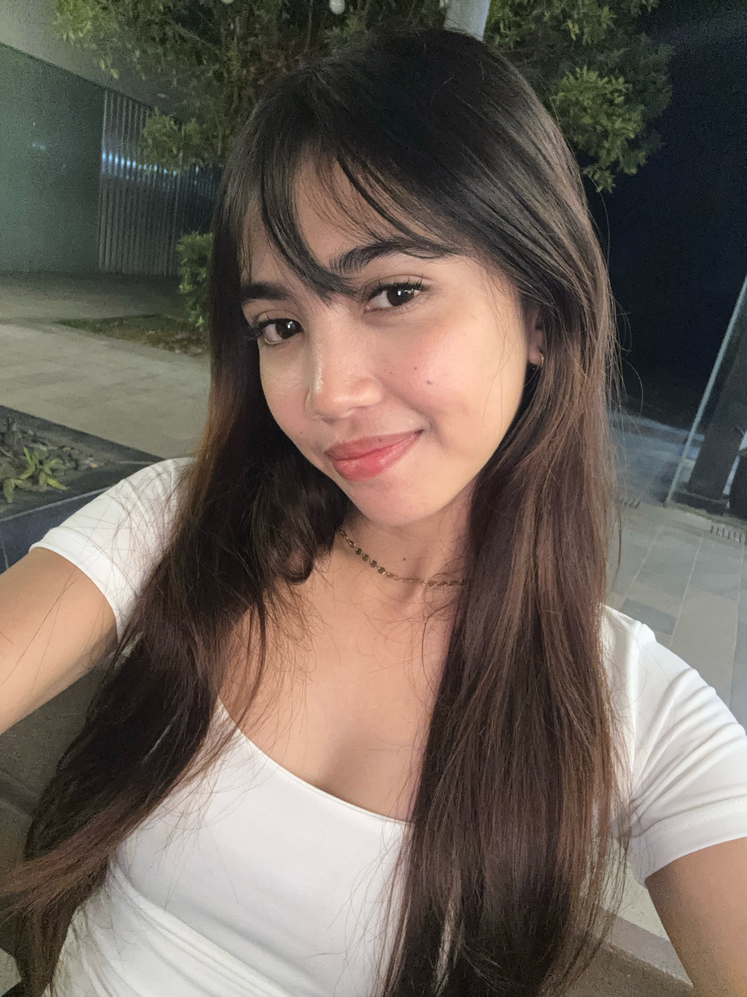 A young woman with long, dark brown hair and bangs is captured in a close-up selfie. She is wearing a white top and a thin gold necklace. Her facial features, including her eyes, nose, and mouth, are visible. She appears to be outdoors at night, with a blurred background that includes some greenery, a fence, and paved ground. The lighting suggests an ambient urban setting.