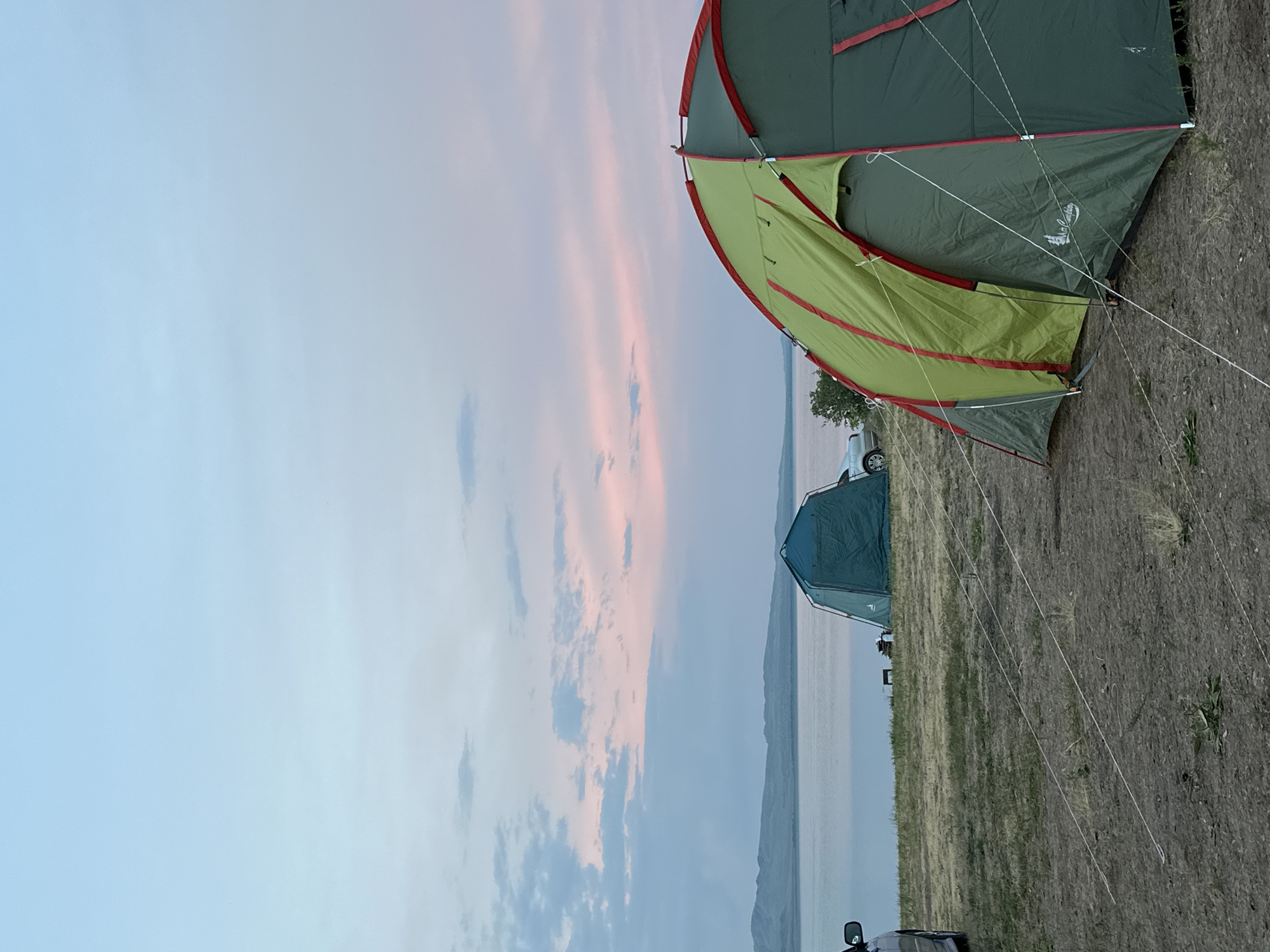 The image depicts a serene campsite at what appears to be either dawn or dusk, given the soft, pastel hues of the sky and the gentle illumination. Two tents are set up on dry, grassy terrain. The foreground is dominated by a large, green and lime-colored tent with red trim, partially open to reveal an inner sleeping compartment. This tent is secured by numerous guy lines, suggesting it is a robust structure designed to withstand elements. In the background, a smaller, dark teal tent is visible, positioned closer to a body of water.
The scene overlooks a vast expanse of water, possibly a lake or a sea, with a low-lying landmass or distant mountains forming the horizon line. The sky is painted with wispy clouds in shades of pale pink, soft blue, and muted gray, indicating a calm atmospheric condition. A silver car is parked near the larger tent, and another vehicle, possibly a black SUV, is partially visible in the lower left corner. The ground around the tents is covered in dry grass and scattered small plants.
The overall mood is peaceful and tranquil, evoking a sense of calm and connection with nature. The time of day suggests a quiet transition between day and night, a popular time for campers to relax and enjoy the scenery. No people are explicitly visible in the image. There is faint text on the larger tent, appearing to be a logo or brand name, possibly including the word "Nordisk" or a similar stylized script, though it's difficult to read clearly due to resolution and angle. The location context of Kolodeznyy, Russia, places this scene within a natural, possibly rural or semi-wild environment.