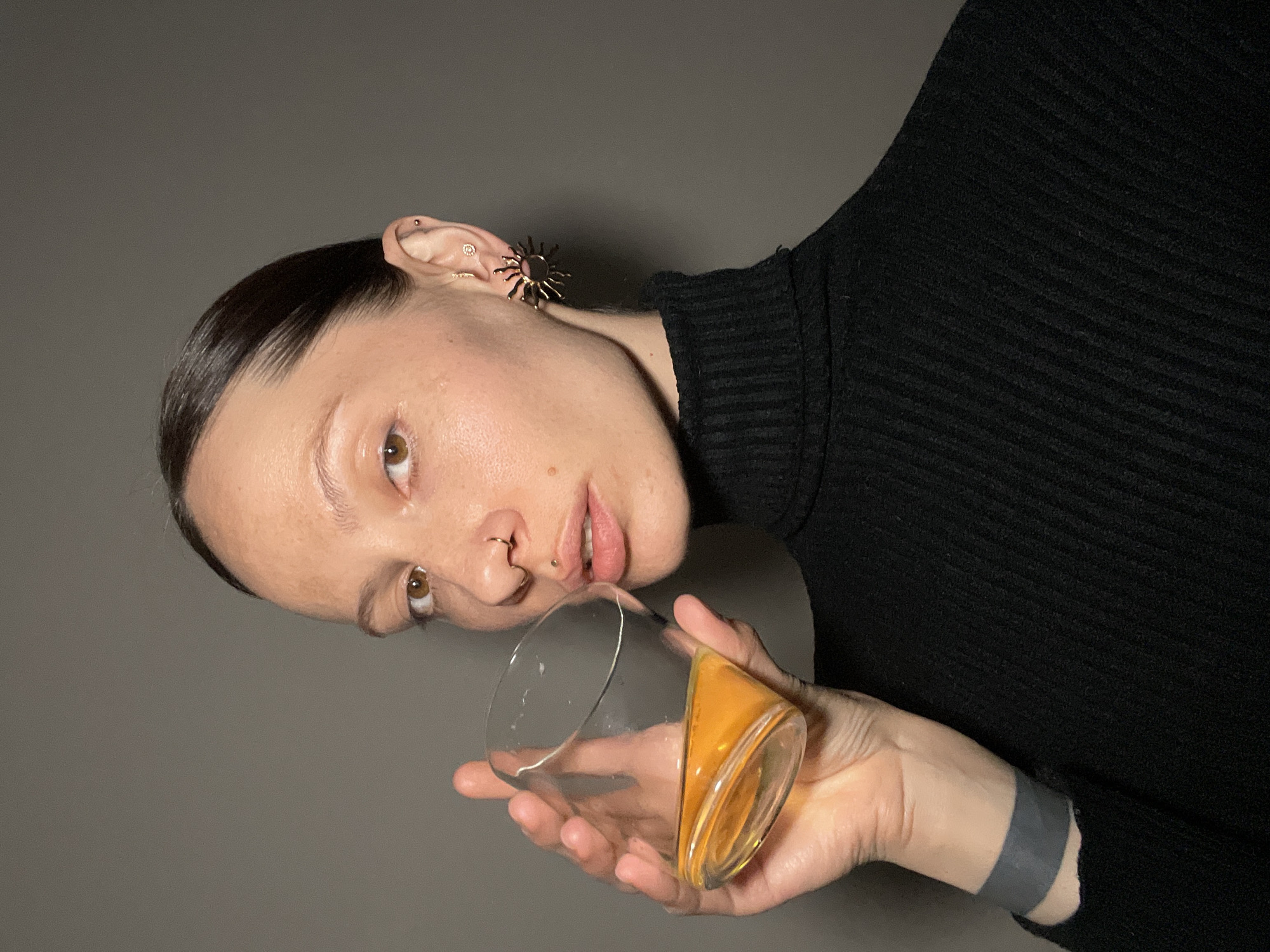 A woman with dark hair pulled back is shown from the chest up, holding a glass with a yellow liquid. She is wearing a black turtleneck sweater and a large, sun-shaped earring. She has piercings in her ear and nose. The background is a solid grey color. There is no indication this image was taken in the city of Pervomaysky, Russia.