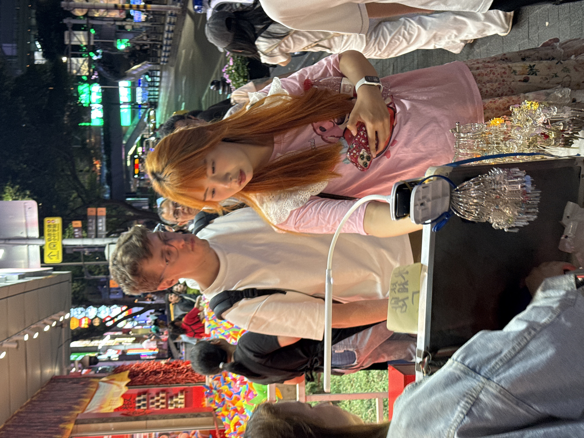 The image captures a bustling street scene in Yuzhong District, China. A young woman with reddish-orange hair, wearing a pink t-shirt with white lace trim and floral-patterned pants, is looking at a display of shiny, metal objects on a black surface. She is holding a phone in her left hand. A person with short blonde hair and glasses, wearing a white t-shirt and a black backpack, stands beside her. A light stick illuminates the display. In the background, there's a vibrant mix of colors and lights, indicative of a busy commercial area, with multiple people walking around. The background lights include green and blue signs. Further back, there are displays of red and other colorful objects. A person wearing a denim jacket is partially visible in the foreground, to the lower right.