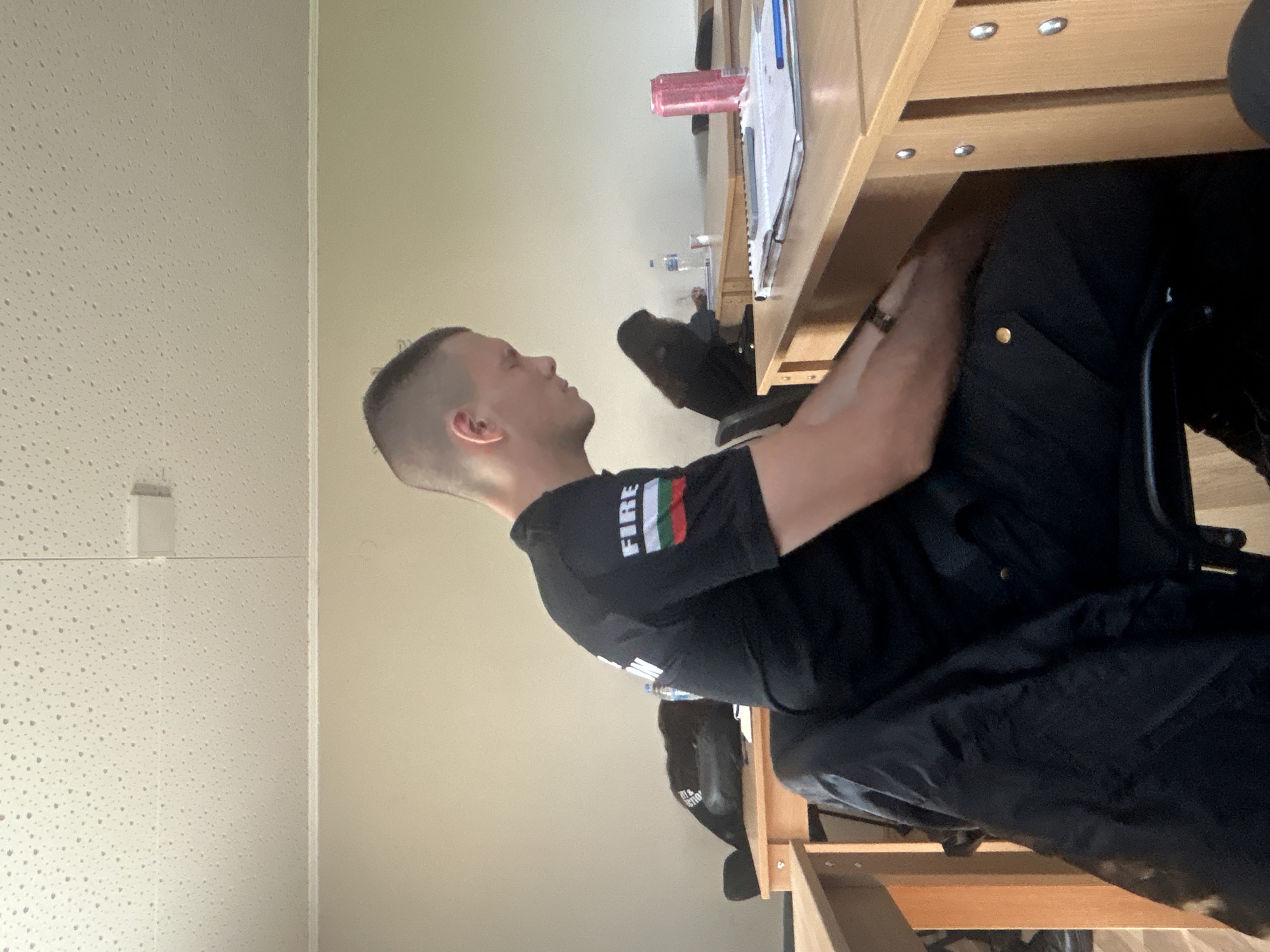 A medium shot captures a man seated at a desk, seemingly in an indoor environment within the city of Unknown, Unknown. The man is positioned to the side, his head tilted slightly upwards. He is wearing a dark-colored t-shirt with the word "FIRE" printed on the sleeve, accompanied by what appears to be a flag or stripe pattern. His hair is short and neatly trimmed.
The desk is made of light-colored wood and contains various items, including a red container, some papers, and a writing utensil. There is also a water bottle in the background. The man's left hand is visible, and he is wearing a ring on one of his fingers. He is dressed in dark clothing.
The background includes a beige wall and a white textured ceiling with a light fixture.