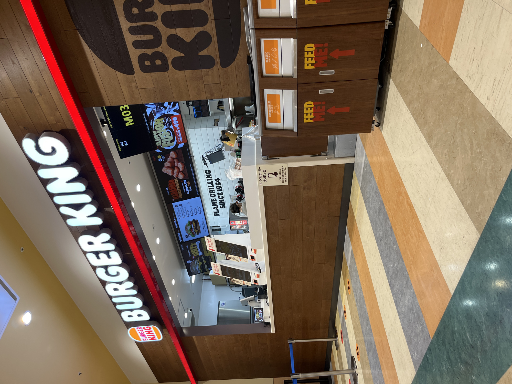 This is a medium-shot, eye-level, exterior photograph of a Burger King restaurant located in Tokorozawa, Japan. The Burger King logo and name are prominently displayed on a wooden-paneled facade. The restaurant features a counter with digital menu boards and self-ordering kiosks. There are also trash receptacles with "FEED ME!" written on them. The floor is covered in a striped pattern of various colors. The scene is brightly lit, suggesting it is daytime.
