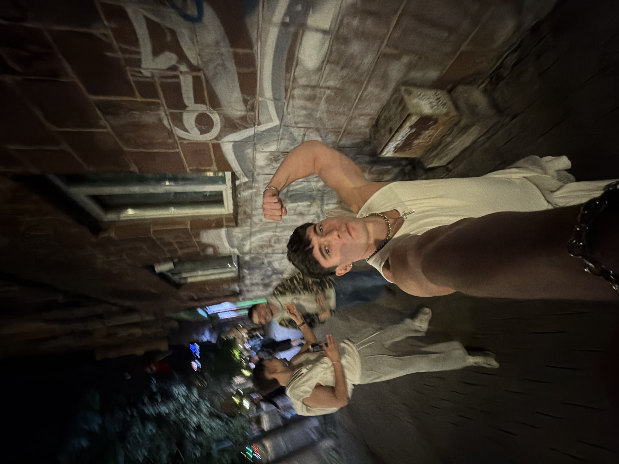 The image shows a selfie of a young man flexing his bicep. Two other people stand behind him. One is holding a camera. The other stands further back in the darkness. The selfie is taken outside on a cobbled street next to a brick building. Graffiti covers the wall of the building. In the distance, the lights of a city can be seen. This scene is likely in Sofia, Bulgaria.