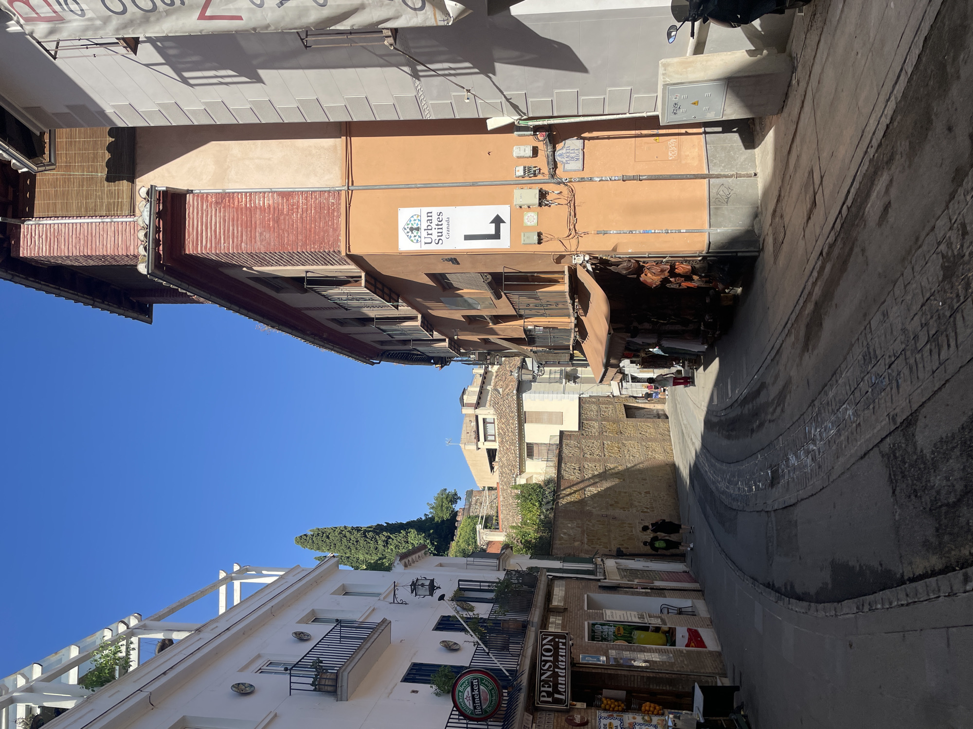 The image depicts a narrow, winding street in Granada, Spain, under a clear blue sky. The street is paved with cobblestones and has tram tracks running down its center. On the left, a white building features a balcony with black railings, windows, and a sign that reads "PENSION Landázuri". Above the pension, a Heineken sign is visible. Further up the street, a tall cypress tree stands against the sky, and the wall of an ancient stone structure is prominent. On the right side of the street, an orange building with multiple balconies dominates the view. A sign on this building reads "Urban Suites Granada" with an arrow pointing left. Several electrical boxes are mounted on the wall. Shops with awnings selling leather goods and other items are located at the base of the orange building. A few people are walking in the street, and a scooter is parked on the right. The architecture suggests a historic European city with a mix of old and renovated buildings.