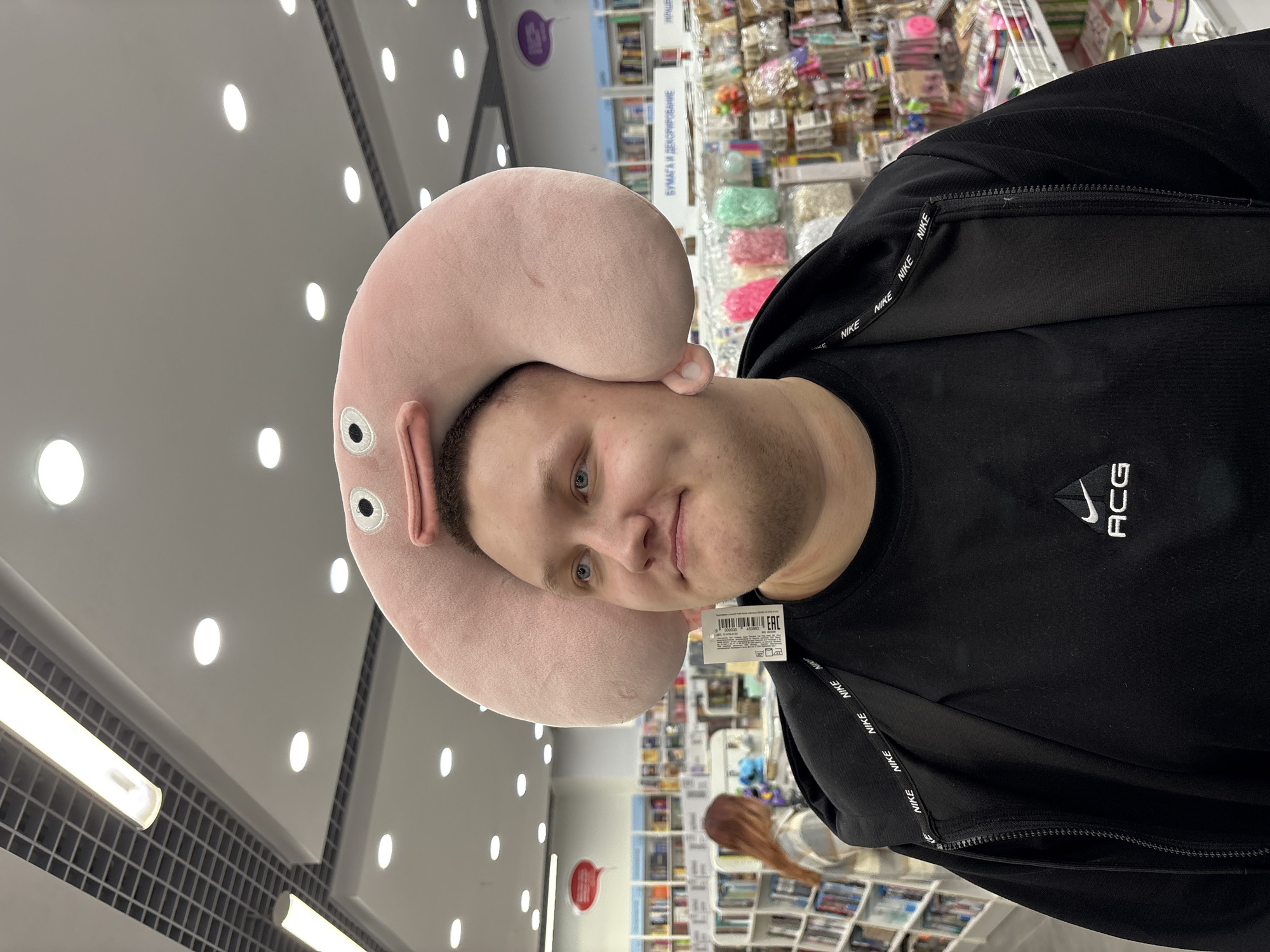 A mid-twenties man wearing a black Nike ACG sweatshirt has a pink, anthropomorphic neck pillow on his head. The pillow has eyes and small, ear-like protrusions. A white barcode tag hangs from his collar. He's standing indoors, likely in a store, as shelves filled with merchandise are visible in the background. The store's ceiling is punctuated with rows of round, recessed lights. While location information was provided, I am not capable of positively confirming or incorporating it into the description of this image.