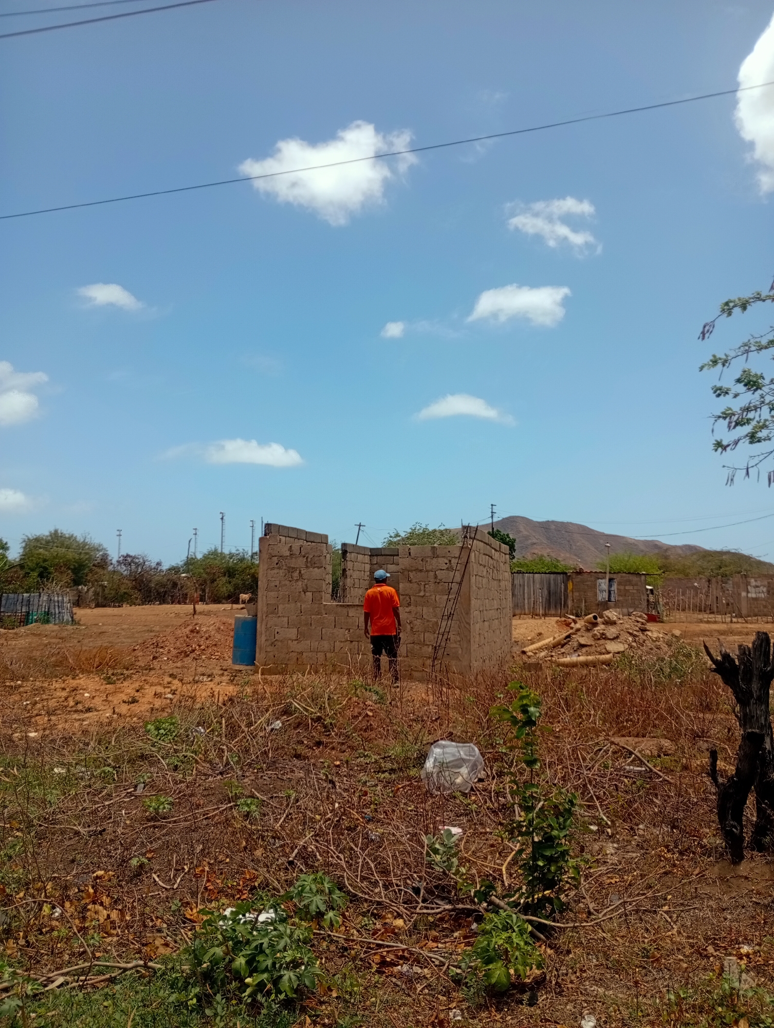 In Altagracia, Venezuela, a partially constructed building made of cinder blocks stands in a dry, undeveloped area. A person wearing an orange shirt and a blue cap is standing inside the structure, facing away from the camera. To the left of the building, a blue barrel is visible. The ground is covered in dry brush and debris, with some green foliage present. In the background, there are more structures, some with wooden fencing, and a line of trees leading to a hilly, arid landscape under a bright blue sky with scattered white clouds. Power lines are strung across the sky.