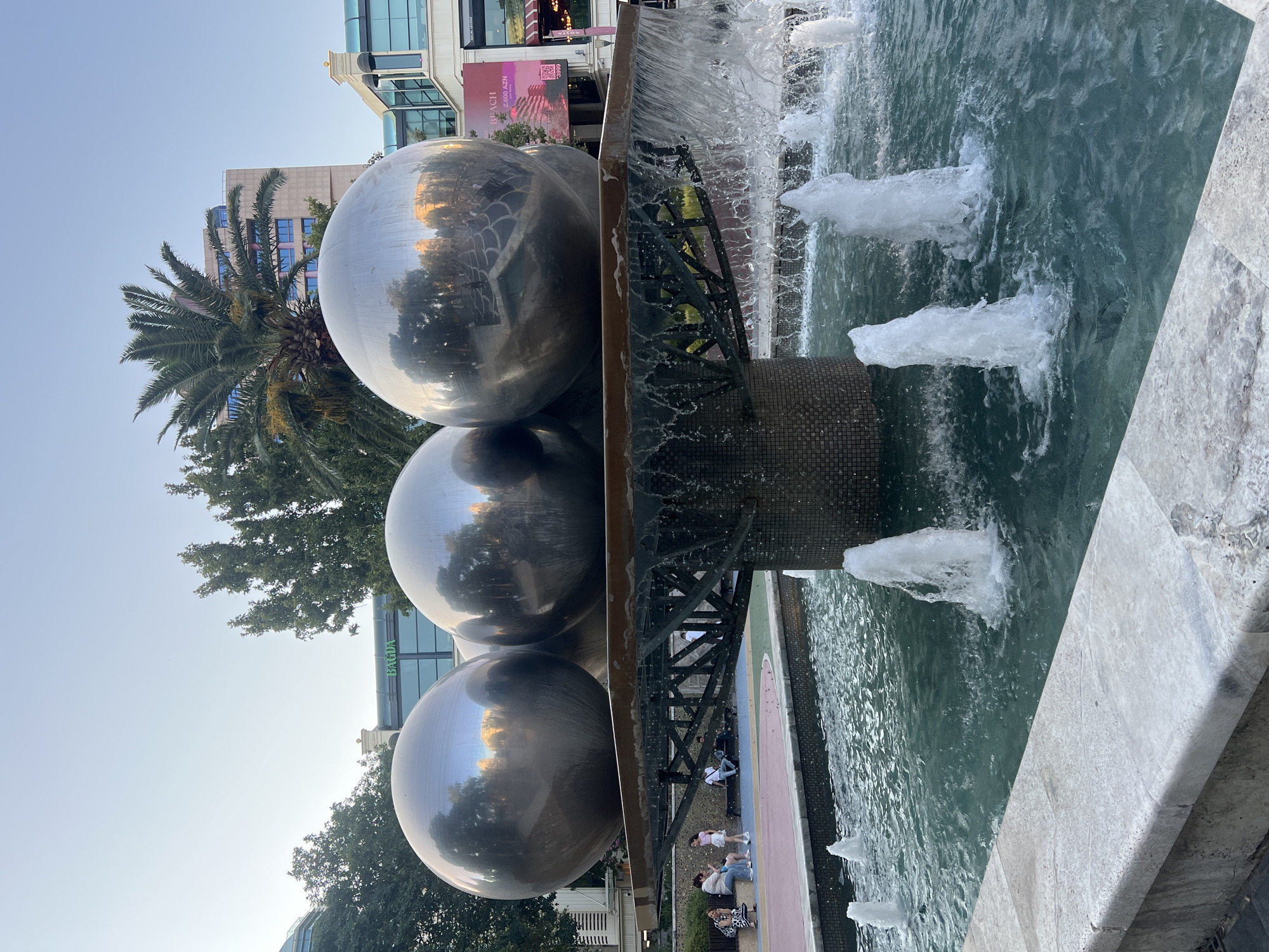 The image shows a public fountain with three large, reflective spherical sculptures at the center. The spheres are made of a polished metal, likely stainless steel, and they reflect the surrounding environment, including buildings and trees. Water cascades from a platform beneath the spheres into a teal-colored pool, where several smaller water jets are active. In the background, there are multi-story buildings, some with glass facades, and lush green trees, including a prominent palm tree. A few people are visible sitting and standing on benches in the middle ground, suggesting a leisurely public space. The paving around the fountain is a combination of light-colored stone and darker patterned tiles. The overall scene depicts a modern urban setting, likely a plaza or park area. Based on visual cues such as the architecture and the presence of the "BAGDA" signage on one of the buildings, the location is likely Sabail Raion, Azerbaijan, possibly in an area with commercial establishments.