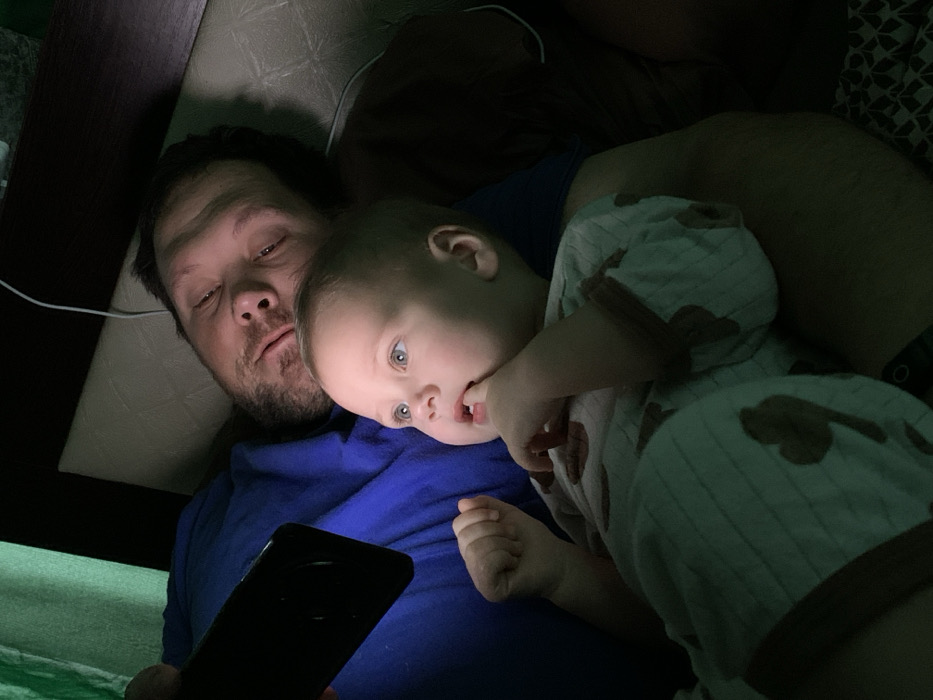 A man and a baby are lying down in bed together. The man is holding a phone, and the baby is leaning on him. The baby is wearing a romper with a brown pattern and has their fingers in their mouth. The man is wearing a blue shirt. The scene is dimly lit, and a charging cable is visible near the man's head.
