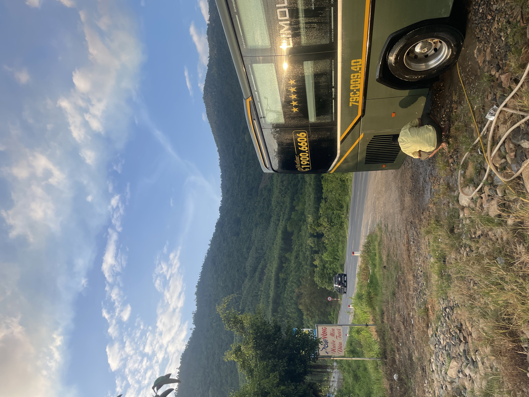 The image depicts a rural or semi-rural roadside scene, stated to be in the city of Tây Ninh Hòa, Vietnam. A large, olive-green "LIMOUSINE" bus with yellow trim and lettering is parked on the right side of an unpaved shoulder. Its side displays "Hotline: 1900.6606", five gold stars, and the license plate "79C-009.40". The bus's side is wet, reflecting bright sunlight. A person, appearing male, is crouched barefoot near the rear wheel of the bus, wearing a light-colored short-sleeved shirt and dark shorts, seemingly engaged in cleaning or maintenance, with several hoses visible on the rocky and grassy ground around him.
To the left, a two-lane paved road recedes into the distance, with a black SUV visible driving away. A circular white speed limit sign with a red border indicating "60" is positioned beside the road. Adjacent to the road, on the left, a white sign with Vietnamese text advertises "Cafe Võng" (Hammock Cafe), "Nước Mía" (Sugarcane Juice), and "Dừa Tươi" (Fresh Coconut). The background is dominated by dense, forested mountains under a clear blue sky with scattered white clouds, suggesting either early morning or late afternoon light, with the sun low on the horizon.