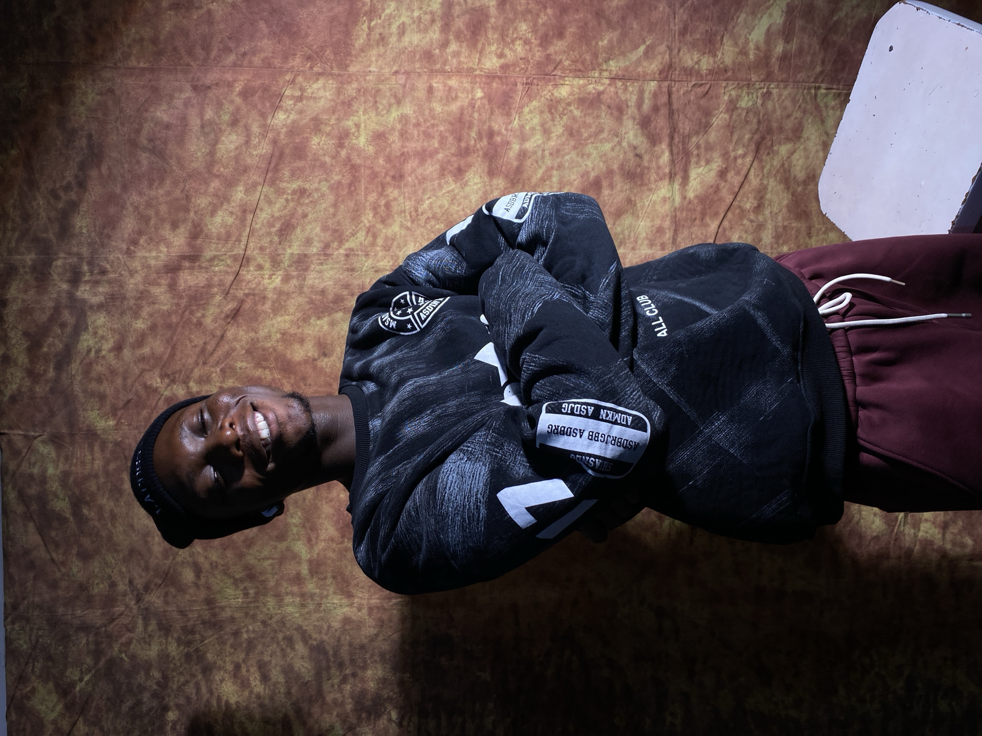 A medium shot shows a Black man wearing a black beanie, a black graphic sweatshirt, and burgundy sweatpants. He stands with his arms crossed and his eyes closed, smiling. The man is positioned in front of a mottled brown and yellow backdrop. The man is wearing blue earbuds. Text on the sweatshirt reads "ALL CLUB". There is a white stool to the right of the frame. The lighting suggests it is daytime. This scene is not in the city of Barakin Akawo, Nigeria.