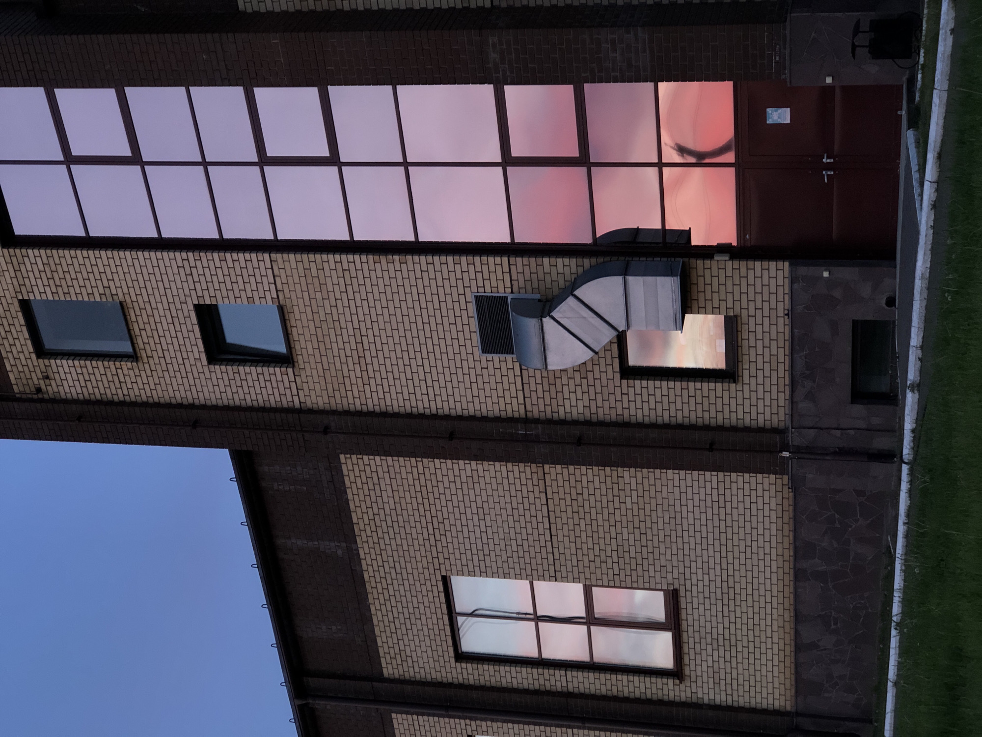 A rotated eye-level view of the exterior of a brick building in Noviy, Russia is depicted at dusk. The lower half of the building is primarily yellow brick with brown brick accents and horizontal rows of darker, almost black brick. The top is a lighter yellow brick with a large window grid that reflects a pink sunset. Two smaller square windows are visible, both reflecting blue dusk light. An industrial air vent system extends from the brick wall next to another window that reflects the pink sunset. A dark red door and a narrow strip of grass are visible to the right. The building is facing left, with a blue sky visible at the bottom left corner.