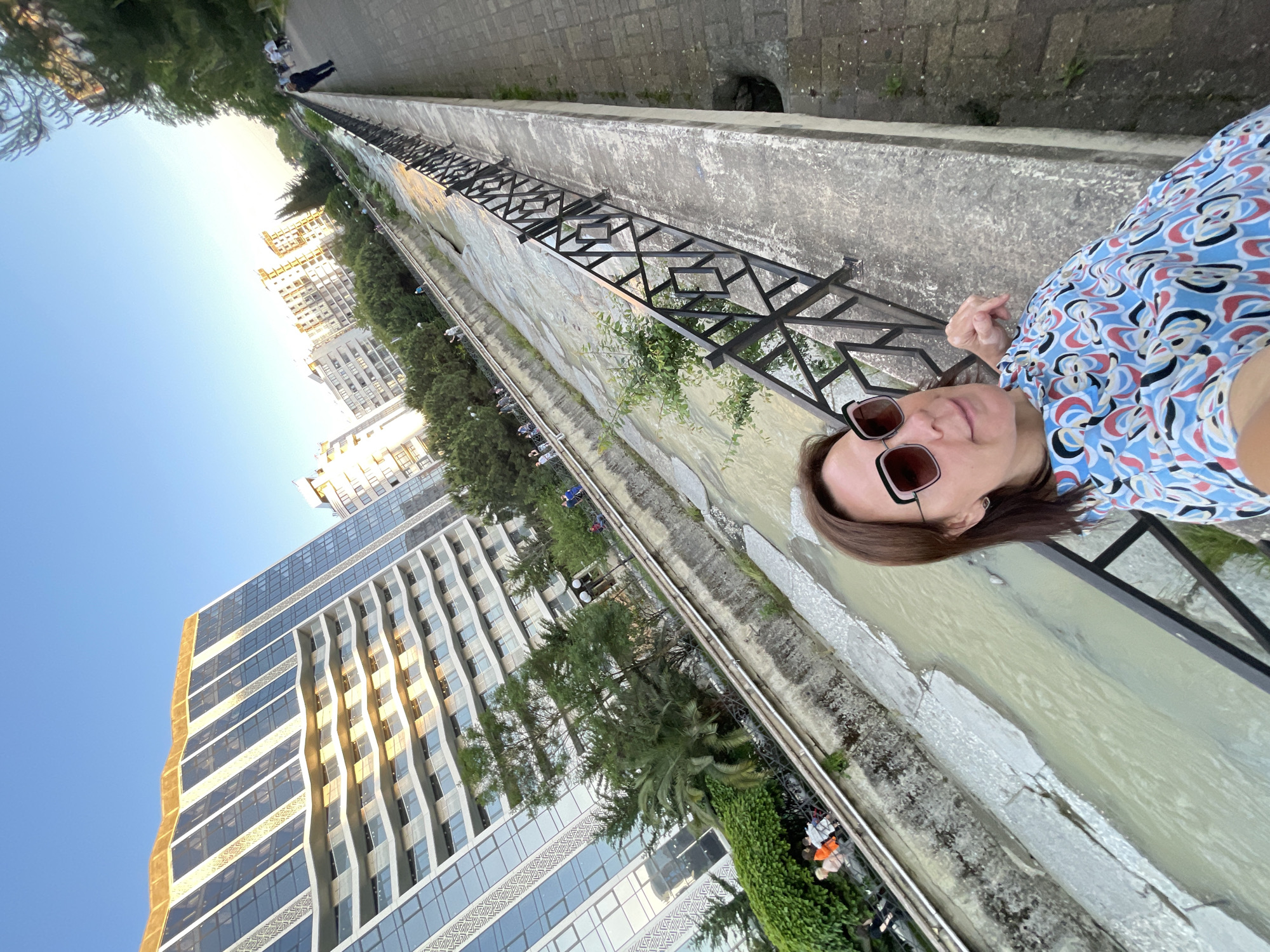 The image is a low-angle selfie, significantly tilted counter-clockwise, depicting a middle-aged woman in the lower right foreground against the backdrop of a concrete river channel and an urban resort environment identified as Sochi, Russia.
The woman has medium-length brown hair and wears dark-lensed, rectangular sunglasses with a thin lighter frame border. She is dressed in a light blue top with a vibrant, multi-colored abstract pattern in shades of red, black, white, yellow, and various blues. Her right arm is visible, holding the camera, and her left hand rests on a black metal railing with a geometric lattice design. She has a slight smile.
Behind her, a wide, shallow concrete channel with murky, light brown water flows from the right to the left. Sparse green bushes and small trees grow from the concrete sides of the channel. A paved promenade is visible on the far side of the channel, lined with trees and occupied by several indistinct figures walking.
In the background, on the left, a prominent modern high-rise building stands, identifiable as the Swissôtel Resort Sochi Kamelia. This structure features a facade of glass and light-colored panels, wavy horizontal balconies, vertical patterned stripes, and a distinctive irregular golden-colored roofline. The building reflects the clear, light blue sky.
On the right side of the channel, parallel to the woman, is a paved walkway made of interlocking grey concrete pavers. A line of mature trees separates this path from several more distant, light-colored multi-story buildings. A person in a dark jacket and trousers is walking away from the camera on this right-side promenade. The scene is illuminated by the warm, low light of a late afternoon or early evening, casting soft shadows. The sky is clear blue, fading slightly towards the horizon.