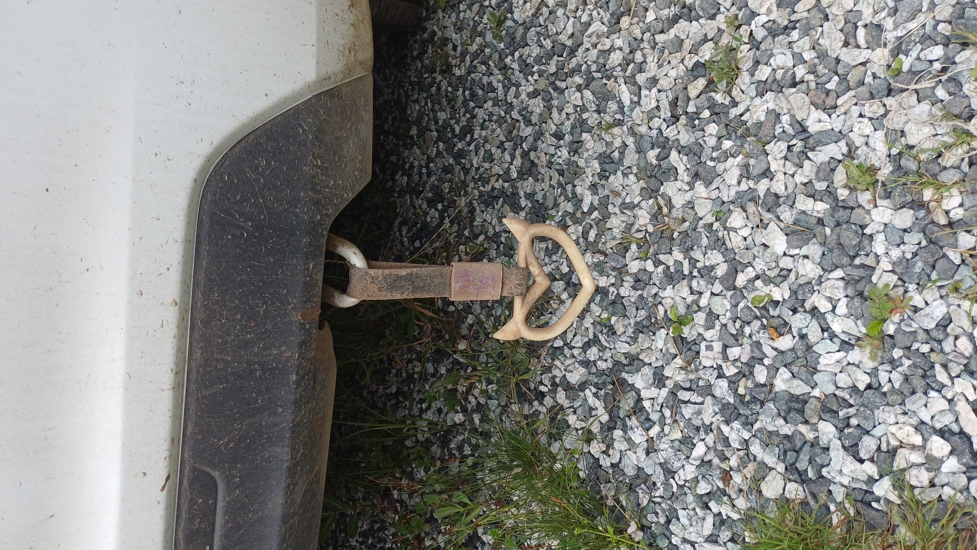 A close-up, eye-level shot shows a decorative tow hook on the rear of a white vehicle. The tow hook is shaped like a heart with two small pointed protrusions on the top outer edges, resembling devil horns. It is a light tan or beige color and is attached to a loop on the underside of the vehicle via a dark, weathered fabric strap. The vehicle's undercarriage and bumper are visible, covered in dirt and dust. The ground below is covered in a mixture of grey and white gravel, with patches of green grass and small weeds interspersed. The scene appears to be outdoors, possibly in a gravel lot or driveway, in Izhevsk, Russia. No people are present in the image.