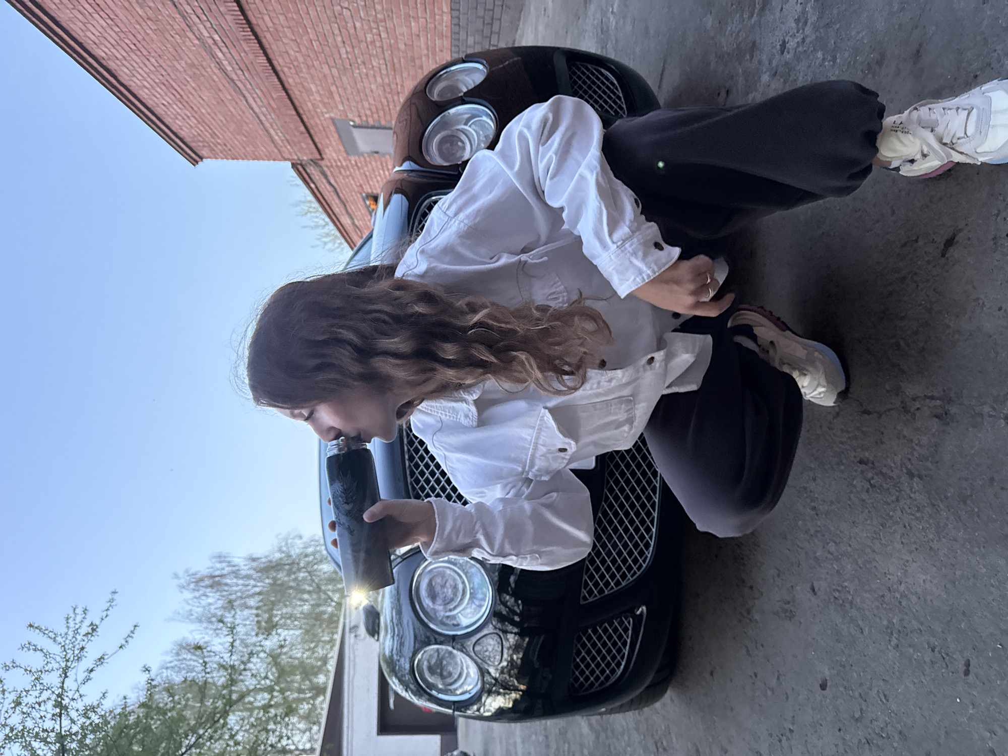 A photograph depicts a young woman kneeling in front of a black Bentley car. She is drinking from a dark-colored water bottle. The woman wears a white jacket and dark trousers, with one foot sporting a white sneaker and the other a tan sneaker. The background includes a brick building and trees. The image was taken in Chaikovsky, Russia.