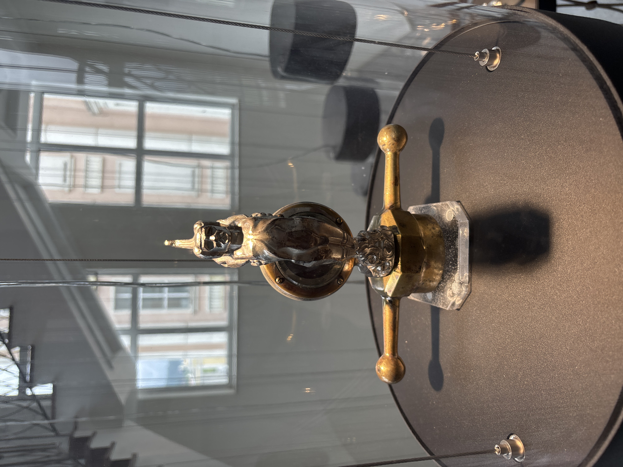 An ornate, silver, mythical creature figurine is displayed behind glass on a black circular pedestal in a museum. The figurine depicts a winged, humanoid figure with exaggerated facial features and a headdress, perched atop a circular metallic base with a lever. The background through the glass shows a blurred view of buildings with windows and a staircase, suggesting an urban or architectural setting. The lighting is focused on the figurine, creating a shadow behind it. The overall environment appears to be an indoor exhibition space. This artifact is located at the Museum of Military Technology in Verkhnyaya Pyshma, Russia.