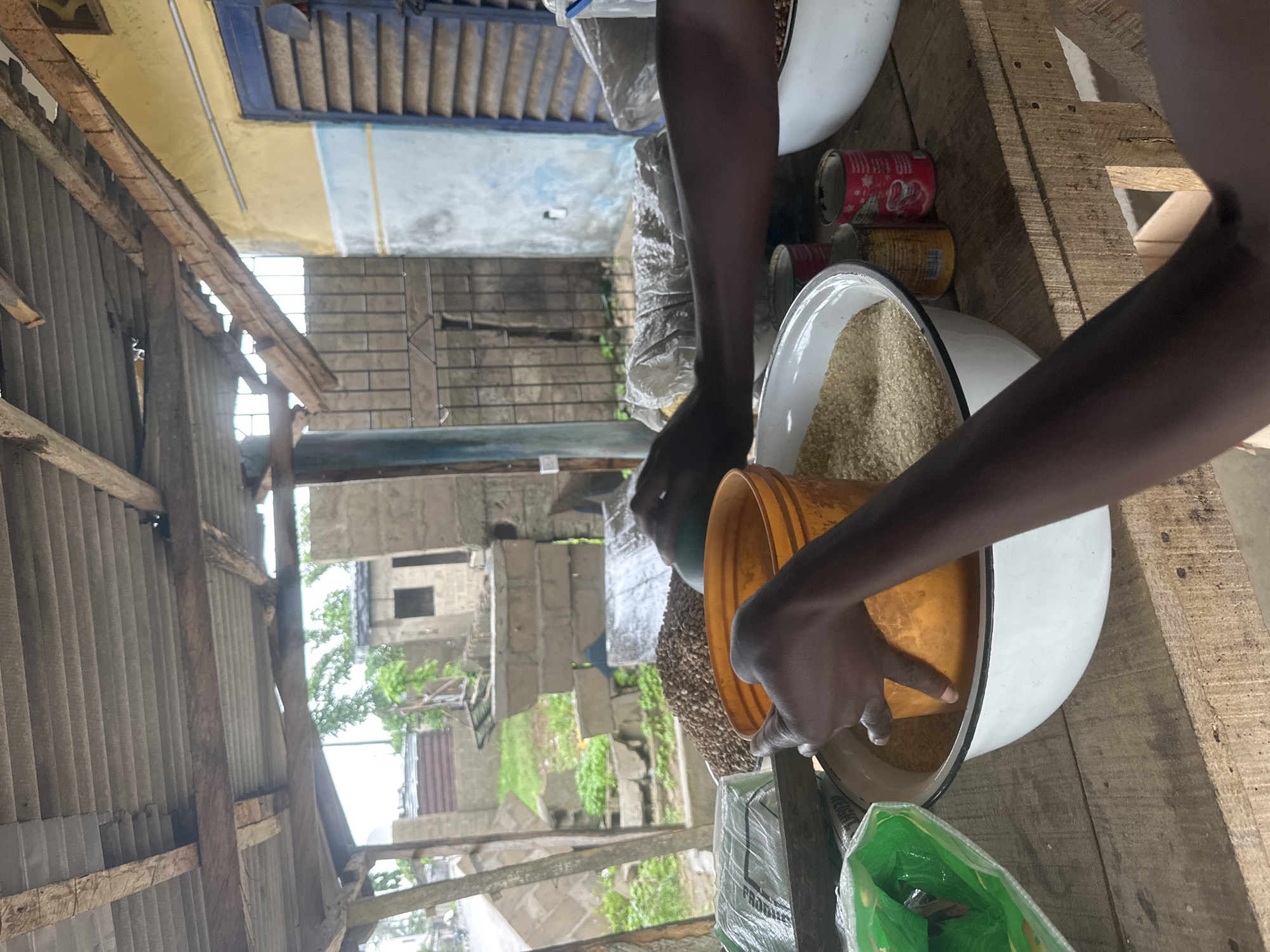 The image shows a close-up view of hands scooping grain from a large white bowl into an orange bucket. The scene appears to be an outdoor market or a food stall, with wooden planks forming the surface where the bowls and cans are placed. In the background, there are buildings constructed with concrete blocks, some of which appear unfinished, and greenery suggesting an outdoor setting. The sky is overcast, indicating it might be a cloudy day.
The subjects are a person's hands, which are dark-skinned, actively engaged in the task of transferring grains. The objects include two large white bowls, one filled with light-brown granular material that looks like rice, and another partially visible, also containing grains. There's also an orange bucket and several tin cans of various sizes, some with red labels. A green plastic bag is visible in the foreground. The setting is rustic, with weathered wooden structures and corrugated metal roofing partially visible, suggesting a simple, perhaps temporary, structure common in market settings.
The activity is food preparation or distribution, specifically measuring or scooping grains. The lighting is diffused, consistent with an overcast day. Location cues like the style of construction and the presence of an open-air market suggest a place in Nigeria, with Badagry being specified. There is no visible text in the image that can be clearly read. The overall impression is one of everyday commerce in a community setting.