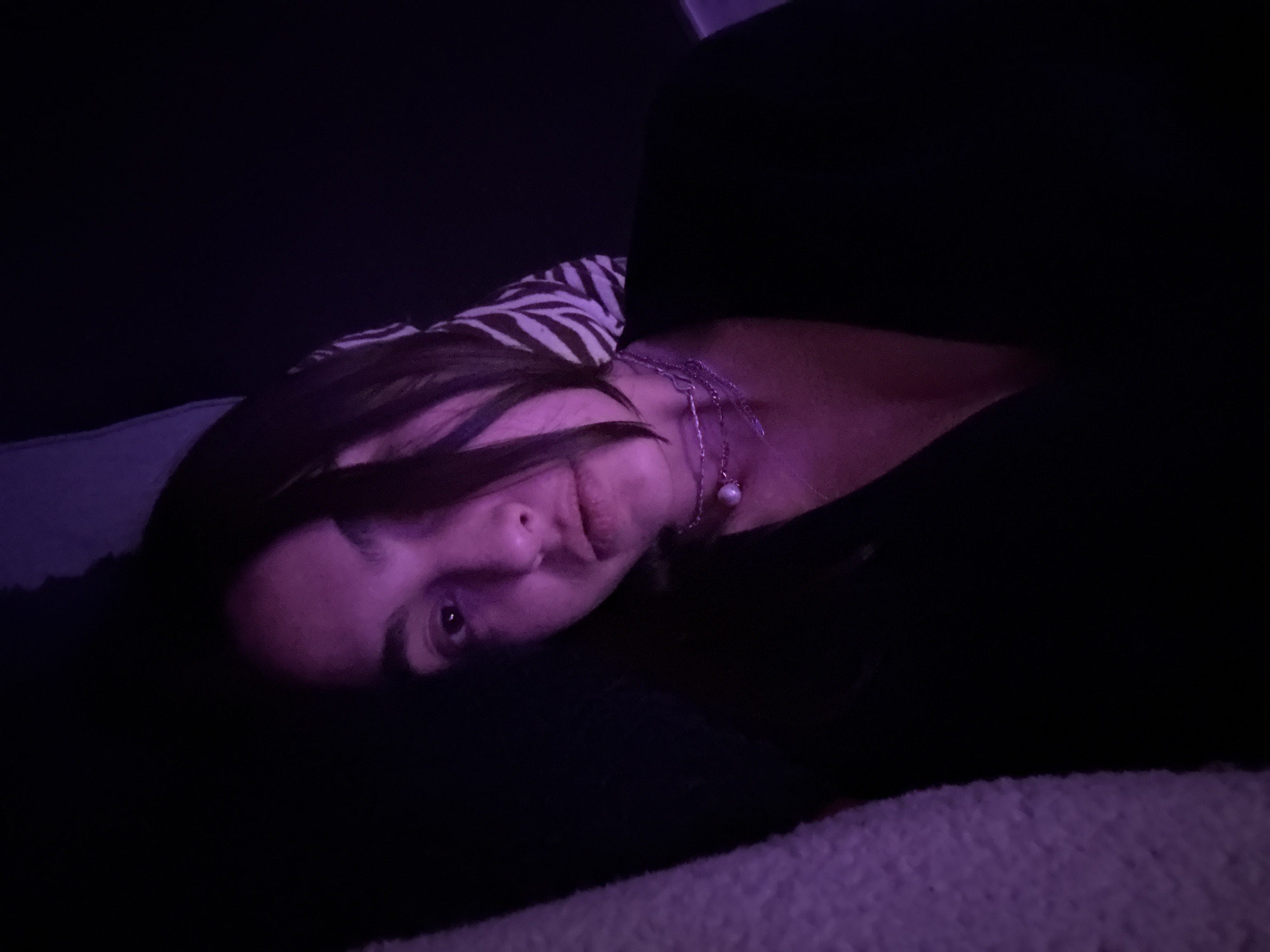 The image displays a young woman resting in a dimly lit, indoor setting, presumably in Mexico City, Mexico. She is lying on her side, with her head resting on a dark surface, possibly a pillow or couch cushion. Her dark hair falls around her face, which is illuminated by a predominant purple-hued light. She has dark eyes and a subtle, closed-mouth smile, looking directly at the viewer.
Around her neck, she wears a delicate silver-toned chain necklace featuring a small, light-colored, spherical pendant, possibly a pearl. Behind her head and shoulder, a patterned fabric with black and white stripes, resembling a zebra print, is partially visible. The foreground shows a textured, soft material, likely a blanket or throw, which also appears purple due to the ambient lighting. The low, purple illumination suggests the photo was taken in the evening or at night, creating a calm and intimate atmosphere. No text is visible in the media file.