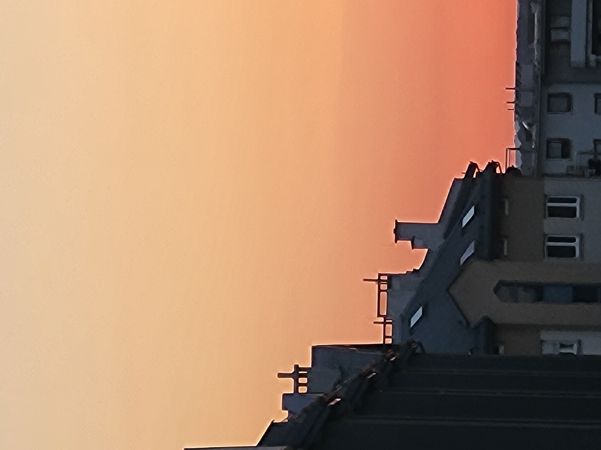 The image depicts a close-up, abstract view of building structures against a warm, gradient sky that transitions from a light peachy-orange to a deeper reddish-orange. The sky suggests either a sunrise or sunset. The buildings are characterized by sharp, geometric lines and appear to be made of concrete and glass. Some sections of the buildings feature rectangular windows, some of which are dark, implying they are either unlit or reflect the sky. The foreground is dominated by dark, angled shapes that could be the roofs or upper levels of these buildings, creating a layered effect. There are no people visible. The overall impression is one of urban architecture set against a dramatic natural sky. The lack of context for the location or any discernible activity leads to an interpretation of the image as a study in form, light, and color.