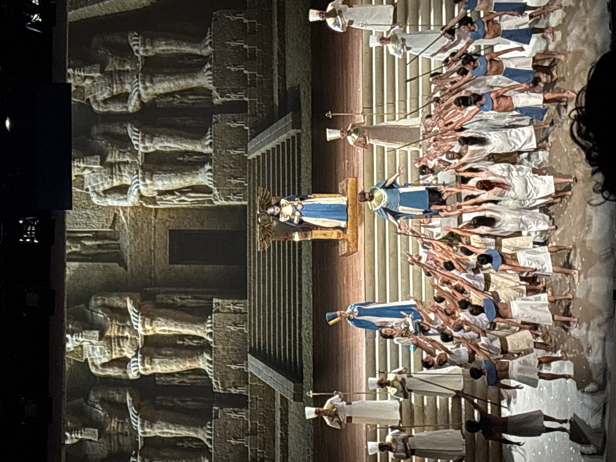 A staged scene of what appears to be an ancient Egyptian opera, displayed indoors. A massive backdrop resembling temple architecture with giant seated figures flanks the left of the frame. Center stage are a pair of figures elevated on a platform, costumed in blue and white. Numerous other figures, similarly costumed, kneel and raise their arms toward the elevated figures. Other figures stand on either side of the kneeling ones, holding what appear to be staffs. The scene suggests a performance or ceremony.
The image does not include information about the location in the city of San Borja, Peru.