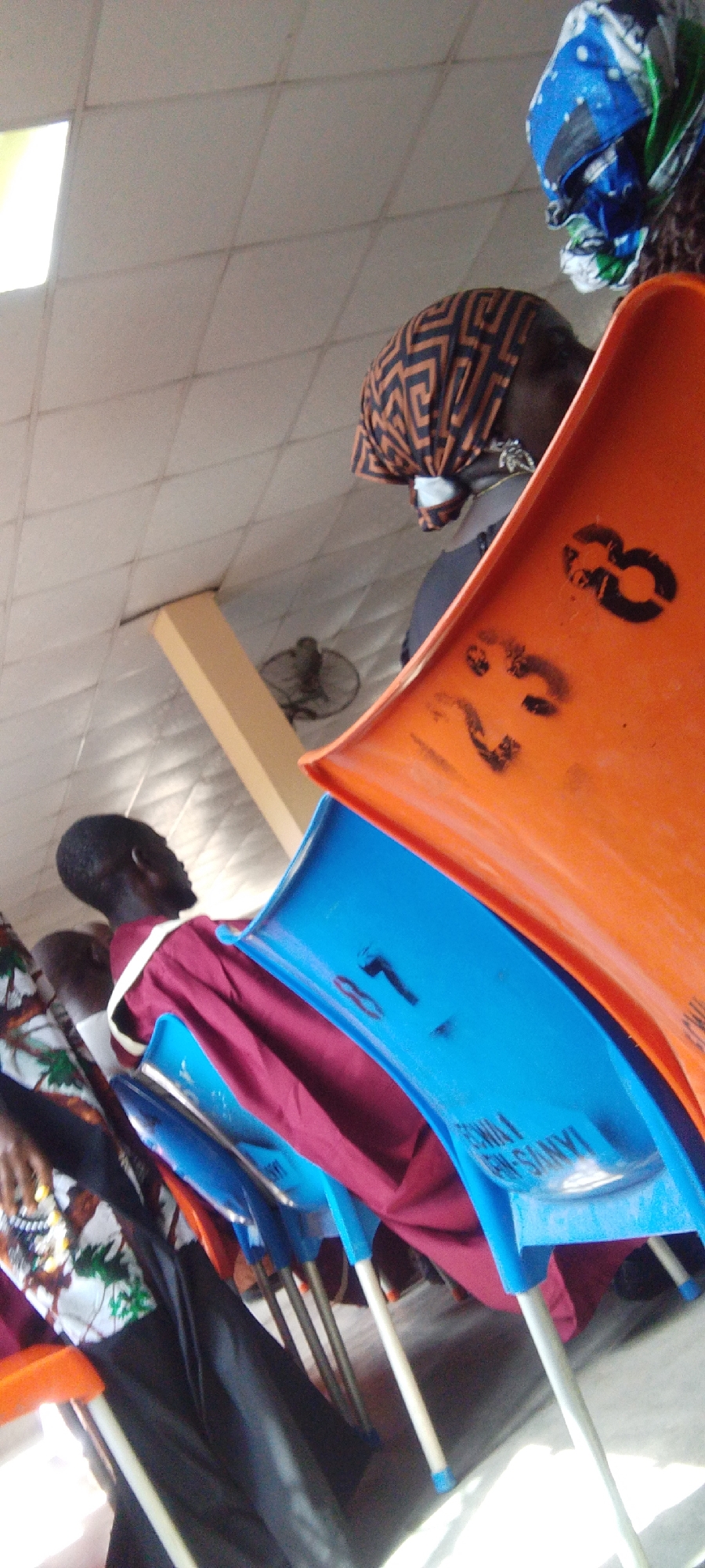 A tilted, medium-shot photograph shows a row of individuals seated on brightly colored plastic chairs in what appears to be an indoor waiting area. The chairs are arranged sequentially with orange and blue seats, each bearing printed numbers and text. The orange chair in the foreground has the numbers "23" and "8" printed on it in black. The blue chair behind it reads "IGIMA1 IAN/VS-MR-SANYI."
Several people are seated in the chairs. One person is wearing a maroon robe with a white sash. Another individual is dressed in a patterned shirt with black pants. Two women wearing headscarves are visible in the background; one with an orange-and-black patterned scarf and the other with a multicolored blue, white, and green scarf.
The environment suggests an indoor setting with a tiled ceiling. In the background, a white column and a ceiling fan can be seen. The image is reported to be taken in Madalla, Nigeria.