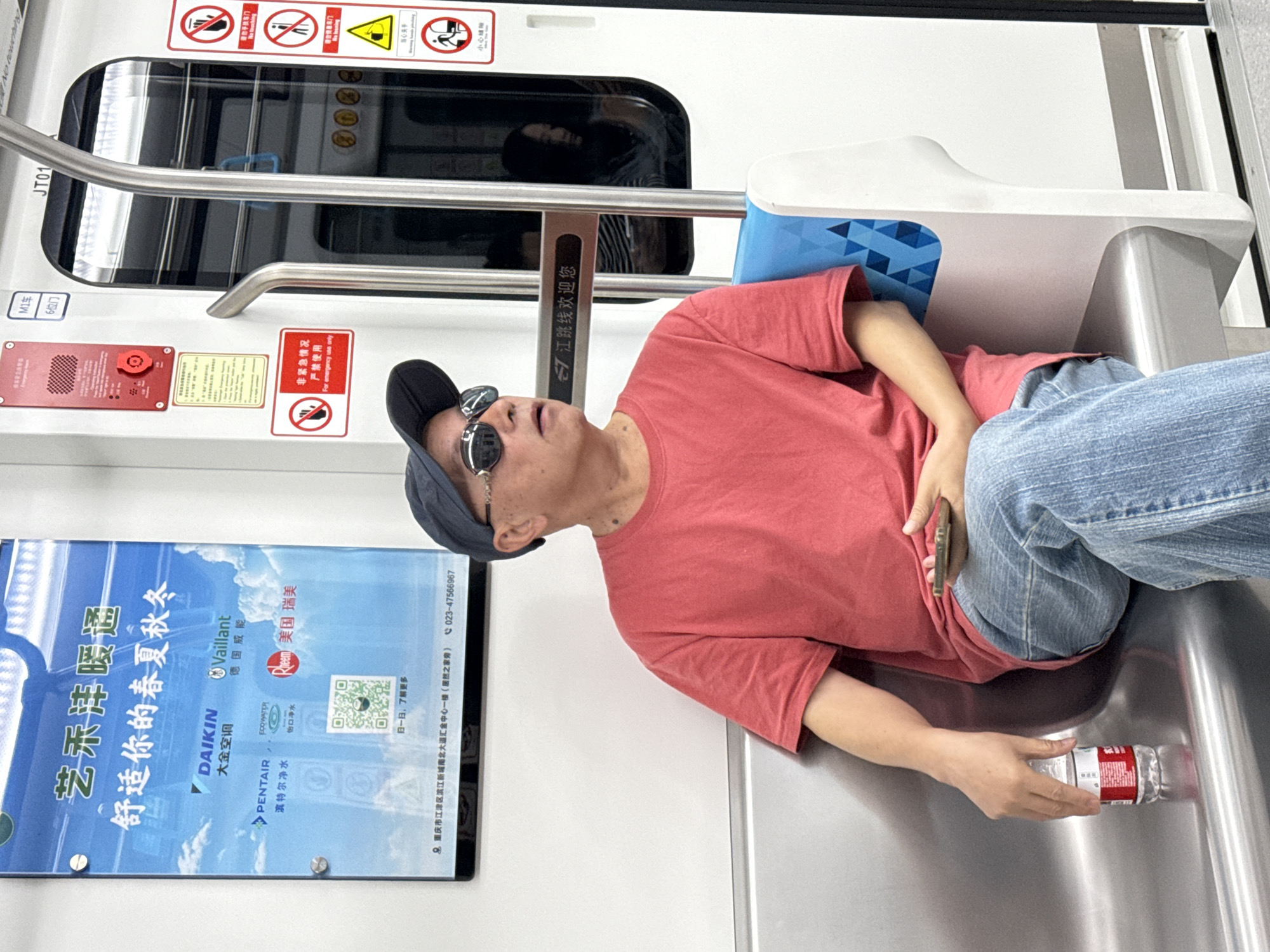 The image depicts a man resting or sleeping across the seats inside a modern subway car, likely in Jiangjin, China. The man is wearing a red t-shirt, blue jeans, a blue baseball cap, and flip-up sunglasses. He is lying stretched out across two silver-colored bench seats, which have blue patterned backs. One of his hands loosely holds a clear plastic water bottle, and a smartphone is tucked into his waist area.
The subway interior features stainless steel surfaces, metal handrails, and windows that reflect the subtle outline of another person. Above the window, several warning signs are displayed. A red sign explicitly states "非紧急情况 严禁使用 For emergency use only" next to an emergency device. Other signs include pictograms and text warning passengers to "当心头手" (Mind your head and hands) and "小心触碰" (Caution, avoid touching). Car identification text such as "JT01," "M1车" (M1 Car), and "6位门" (Door 6) are visible on the wall.
On the left side of the image, a large advertisement board promotes "艺禾洋暖通" (Yihe Yang HVAC) with the slogan "舒适你的春夏秋冬" (Comfortable for your spring, summer, autumn, winter). It features logos for brands like DAIKIN (Daikin Air Conditioning), Vaillant, PENTAIR (Pentair Water Purifier), ECOWATER (Ecobright Water Purifier), and Rheem. The ad includes a QR code, a phone number (023-47566967), and an address in Chongqing Jiangjin District. A smaller sign above the window welcomes passengers to the "江跳线" (Jiangtiao Line). The setting is brightly lit, indicative of an indoor public transit environment.
