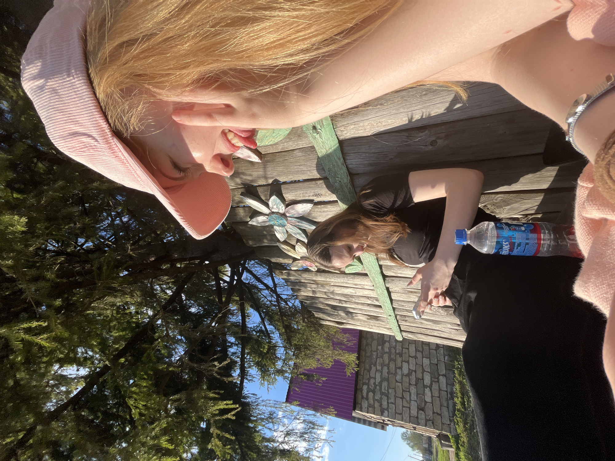 Two females are lying on a wooden surface, likely a picnic table, outdoors. One female is fair-skinned with blonde hair wearing a pink hat. The other female is darker-haired and wearing a black top and pants. There is a water bottle and decorative metal flower on the table. In the background, there are trees, a building with a purple roof, and a stone structure. The scene appears to be in a residential area, with the image possibly taken in Сосново, Russia.