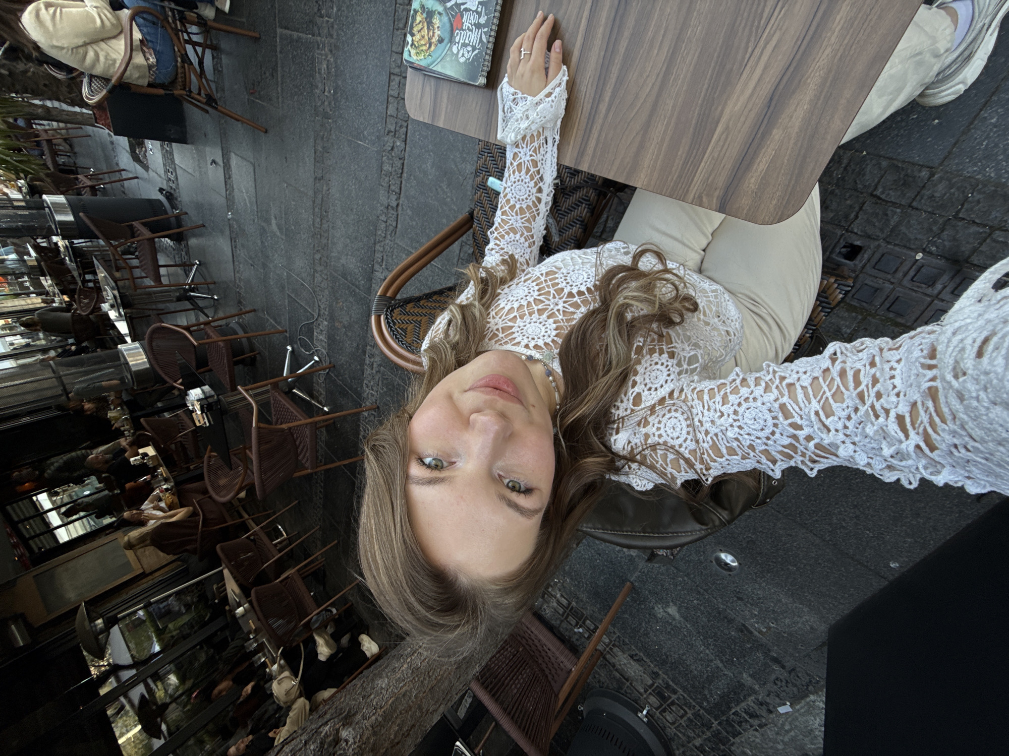 A young woman with long brown hair, wearing a white lace top and light-colored pants, is sitting at a table in an outdoor cafe. She is taking a selfie. A menu is on the table. Other patrons are seated at tables in the background, which include brown wicker chairs. The cafe appears to be in Belgrade, Serbia. The flooring is dark gray pavement. A tree trunk is visible in the lower left corner of the image.