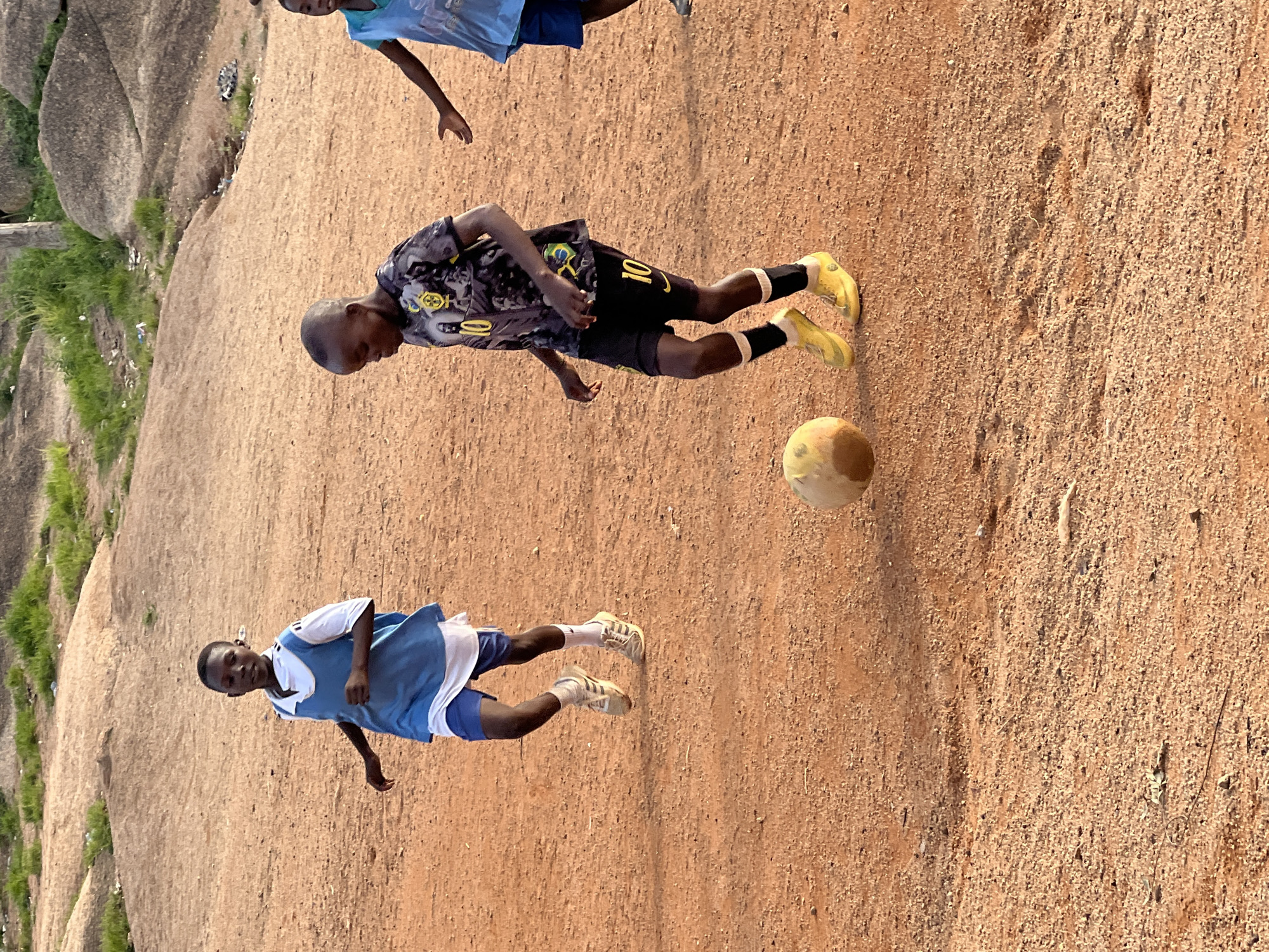 In Jos, Nigeria, three young boys are playing soccer on a dusty, reddish-brown field. The boy in the foreground is wearing a black and grey t-shirt with a graphic print and the number "10" on the sleeve and shorts, paired with bright yellow soccer cleats and black socks. He is dribbling a worn, yellowish soccer ball. To his left, another boy in a light blue jersey over a white t-shirt and blue shorts is running, his feet shod in light-colored sneakers. Further to the right, partially visible, is a third boy in a blue jersey and dark shorts. The background consists of a gently sloping, rocky terrain with sparse green vegetation and some scattered debris. The lighting suggests it is late afternoon.