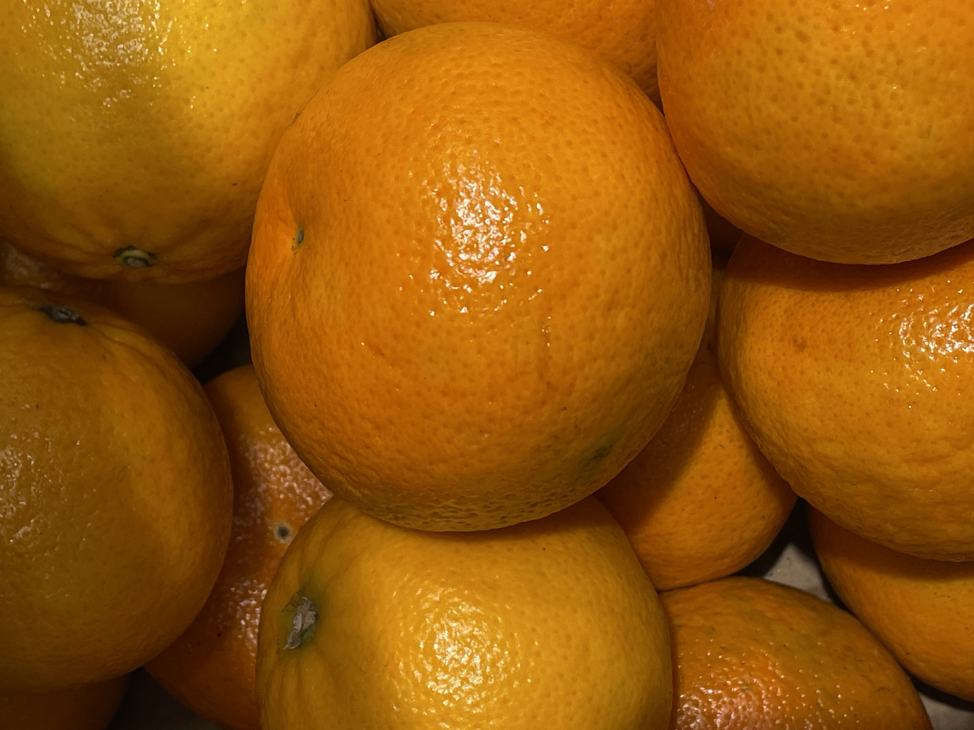 A close-up, overhead shot displays a pile of bright orange tangerines. The tangerines are densely packed, with some overlapping each other. Their textured, bumpy skin is visible, along with small highlights indicating a moist surface. The lighting is warm and appears to come from above, casting subtle shadows between the fruits. The background is a dark, indistinct mass, possibly a container or the surrounding area of a market stall. The image is a still life, focusing solely on the arrangement and visual appeal of the tangerines. There is no indication of people, actions, or interactions. The location in Rostov-on-Don, Russia, is not discernible from the image itself.