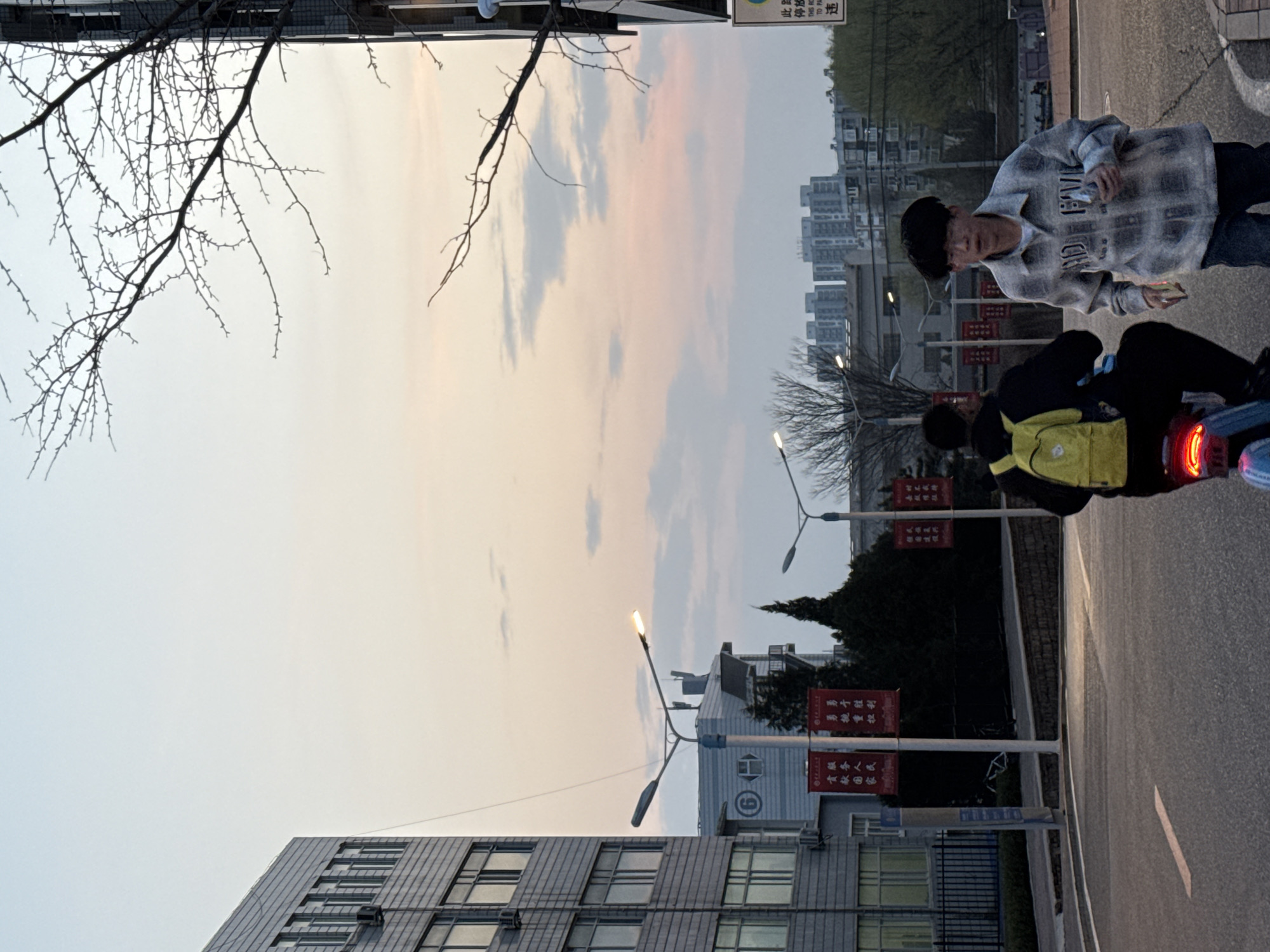 Two individuals are positioned on a scooter on a paved road within Zhongtun, China. The setting is urban with buildings, streetlights, and banners visible. The sky is overcast, suggesting it is either dusk or dawn. One individual is operating the scooter, while the other stands beside it, holding what appears to be a mobile device.