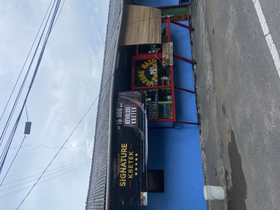 The image shows a small storefront with a blue facade and red trim, located in Cipatat, Indonesia, at coordinates -6.785 latitude and 107.435 longitude. The store has a sign above the entrance that reads "Warung Nasi." The sign is made of wood and has a yellow background with black letters. There is a bamboo blind covering part of the store window. A white bucket is sitting on the sidewalk in front of the store. The store is situated on a paved street with a small patch of grass in the background. Above the store is a corrugated metal roof and there are several power lines running overhead. There is a sign above the store advertising "Signature Kretek" cigarettes with the tagline "Revolusi Kretek". There is a man's face in the top left of the "Signature Kretek" sign. There are several bottles of what appear to be drinks displayed in the storefront window. A white painted white line is on the street in the bottom right corner of the frame. The sky is gray and cloudy. The store is located on a narrow street in a small town, with a small amount of vegetation visible in the distance.