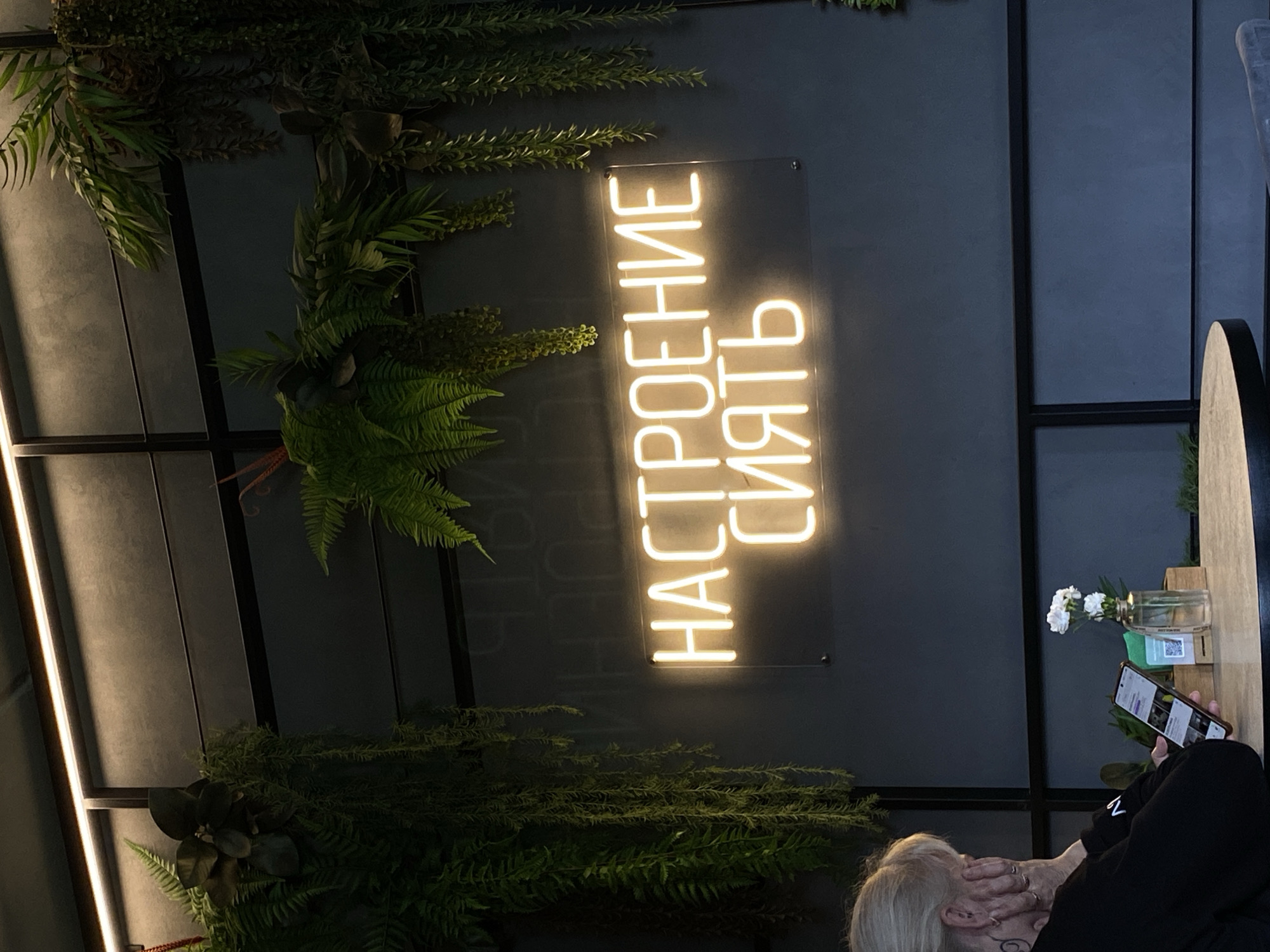 The image shows a dimly lit interior with a neon sign that reads "НАСТРОЕНИЕ СИЯТЬ" (Mood to Shine) in glowing yellow letters against a dark gray wall. The wall is decorated with artificial green plants hanging from above. In the foreground, a person with blonde hair, seen from the side, is sitting at a table, looking at their phone. The table has a small vase with white flowers and a QR code. The overall atmosphere appears to be in a cafe or restaurant. The location is Votkinsk, Russia.